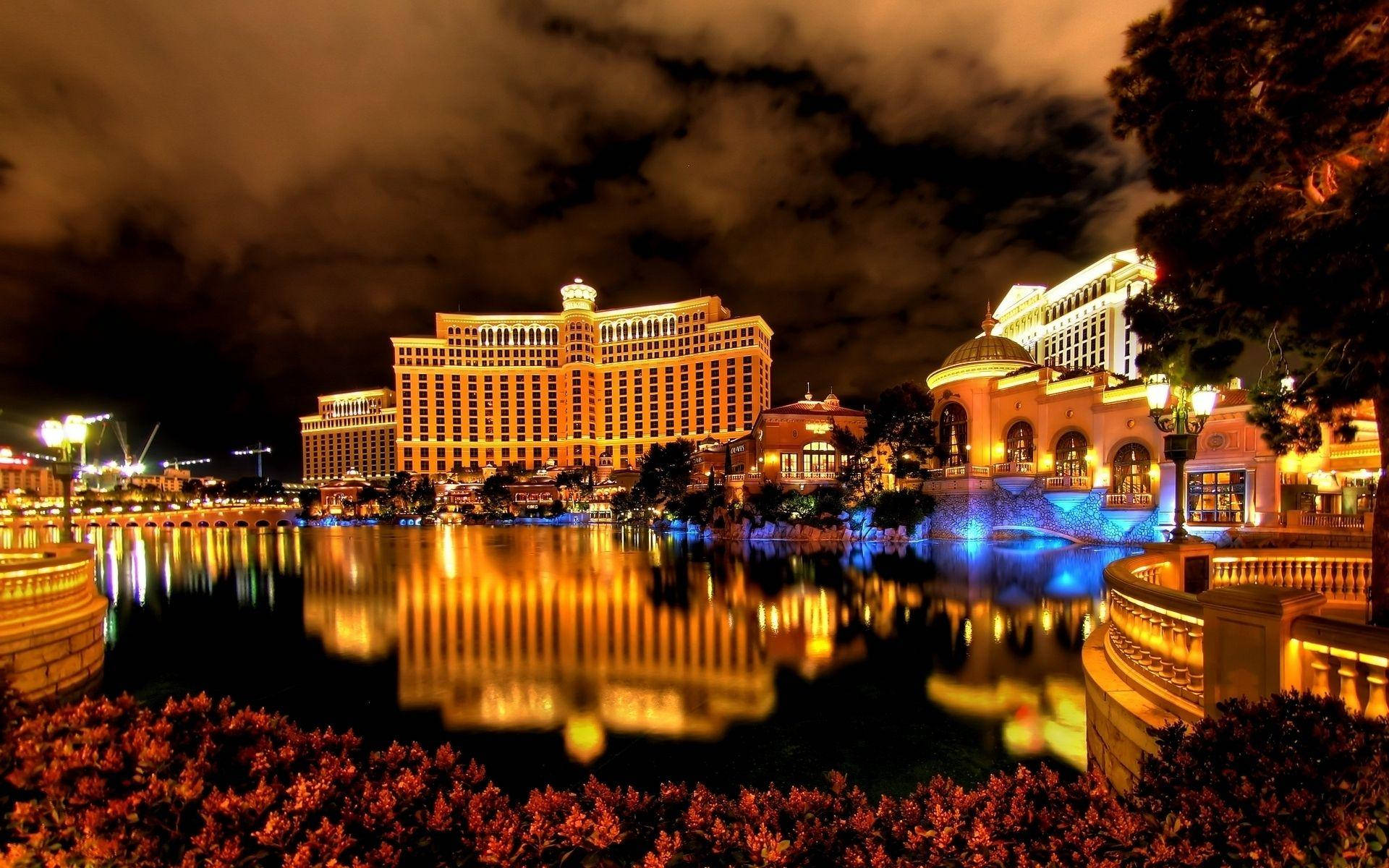 Las Vegas 桌布 壁紙 — 一個大型飯店綜合體在夜晚溫暖地發光，其燈光在多雲的天空下平靜的水景中生動地反射。