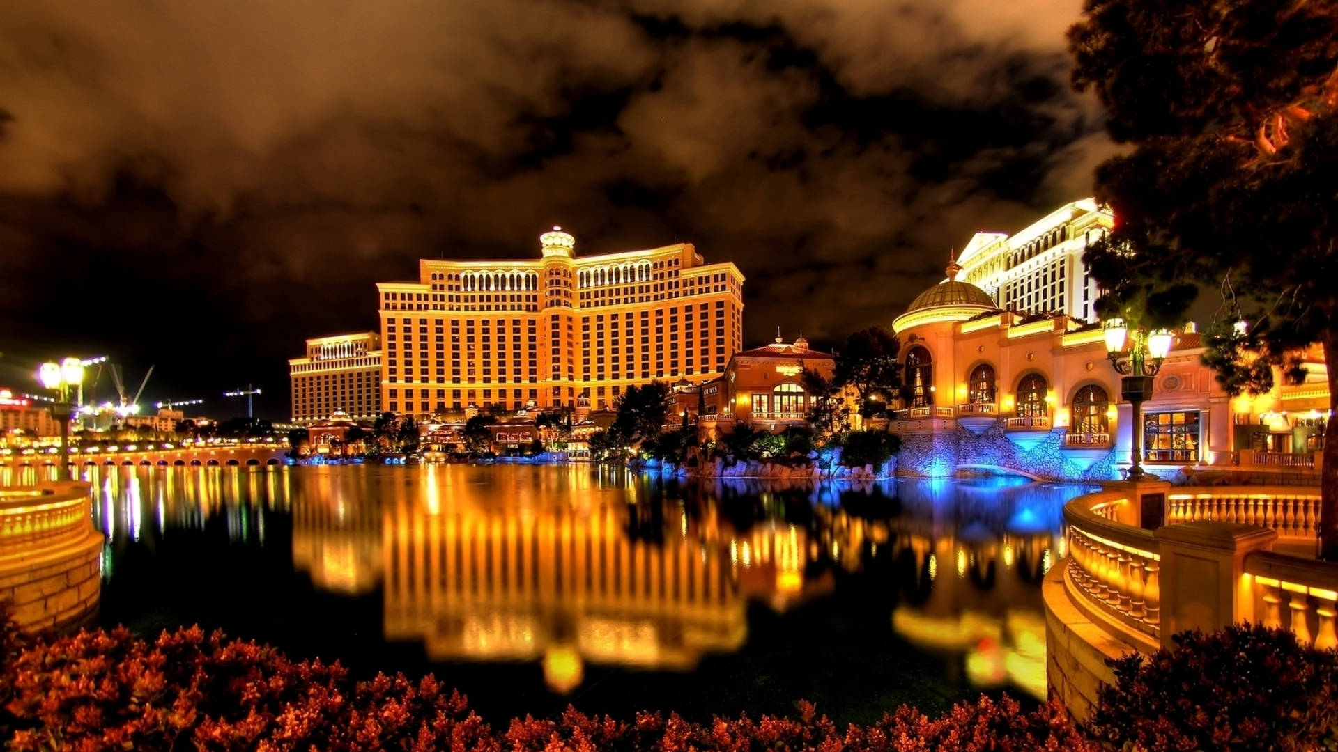 Las Vegas 桌布 壁紙 — 一個大型飯店綜合體在夜晚溫暖地發光，其燈光在多雲的天空下平靜的水景中生動地反射。