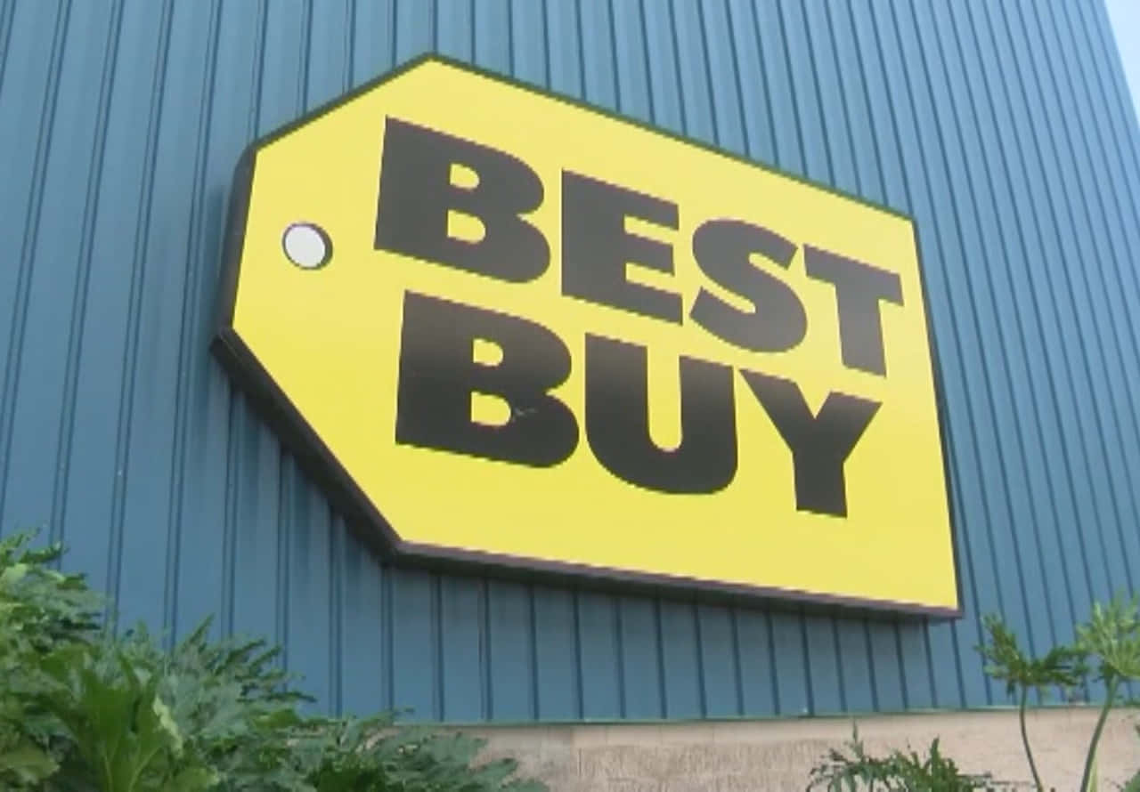 Best Buy 桌布 壁紙 — 一個大型黃色Best Buy價格標牌安裝在藍色波紋金屬建築外牆上，底部有綠色植物。