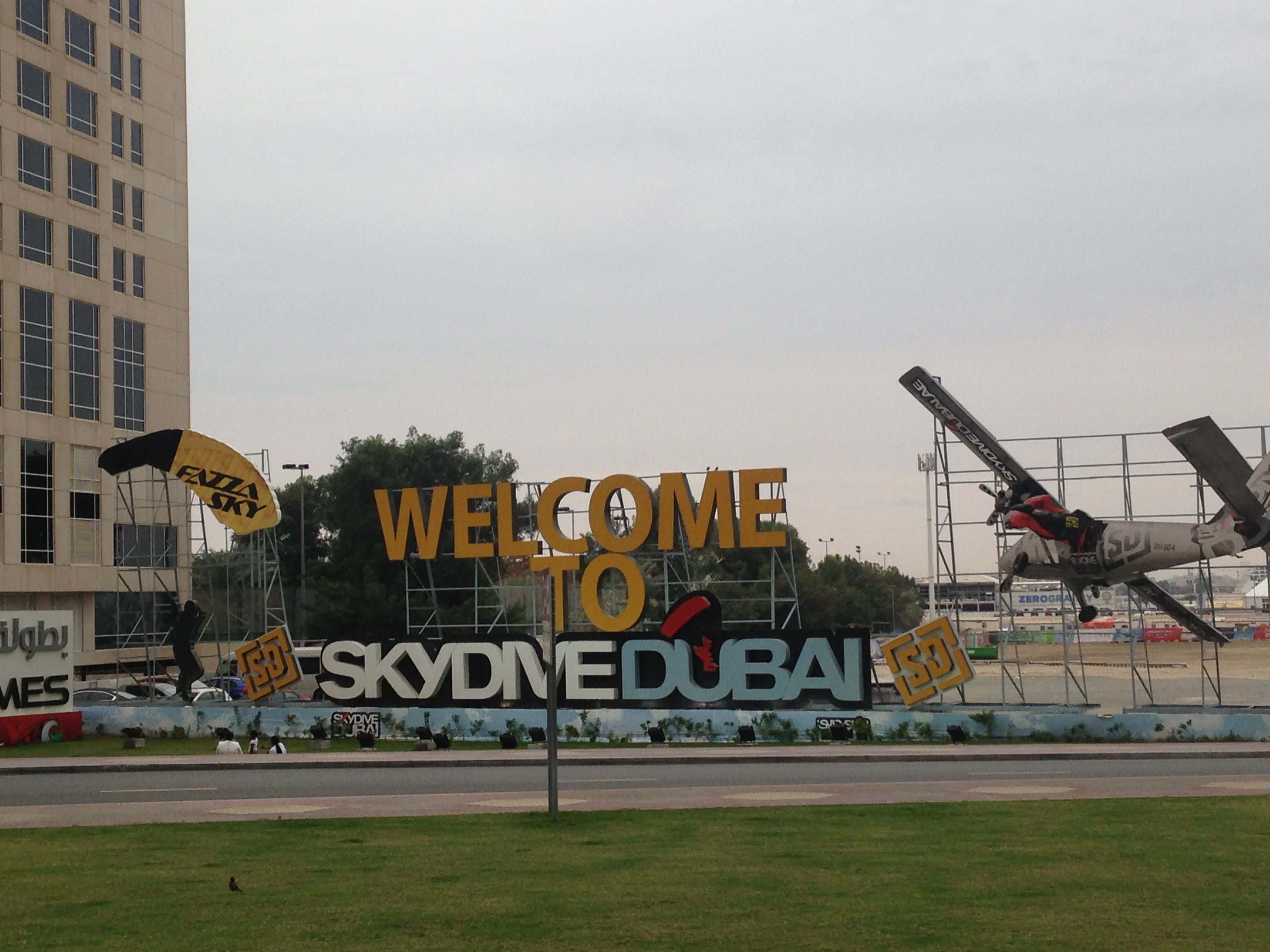 杜拜 桌布 壁紙 — 一個大型黃色標牌，上面寫著「WELCOME TO SKYDIVE DUBAI」，旁邊懸掛著一架飛機和一個降落傘，背景是多雲的天空。