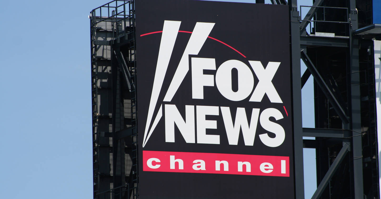 News 桌布 壁紙 — 一個大型黑色Fox News Channel標誌，帶有白色和紅色字體，安裝在金屬結構上，背景是清澈的藍天。