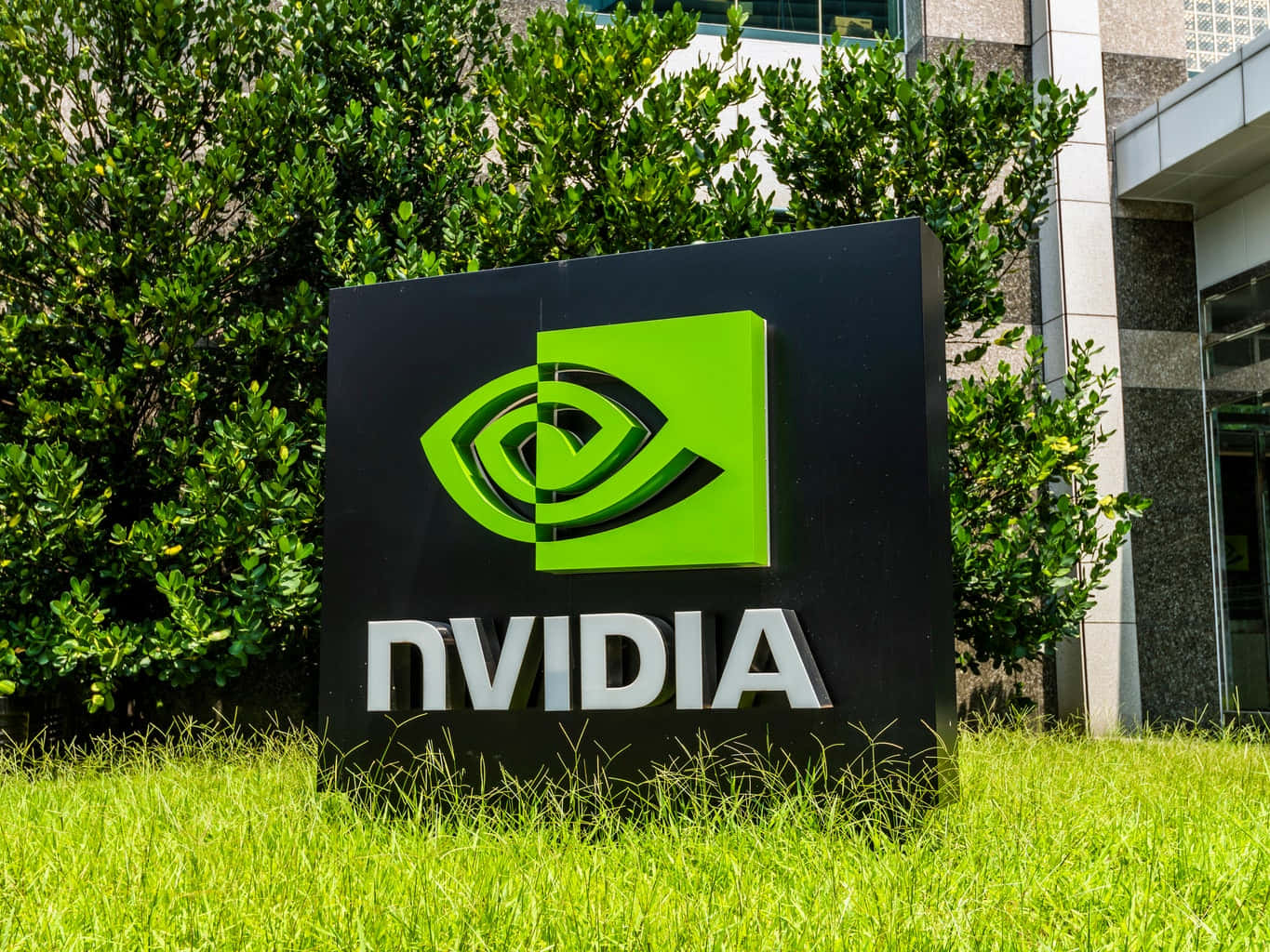 nvidia 桌布 壁紙 — 一個大型黑色NVIDIA標誌，帶有明亮的綠色眼睛標誌，矗立在充滿活力的綠色草地上，旁邊是現代建築和茂密的灌木叢。