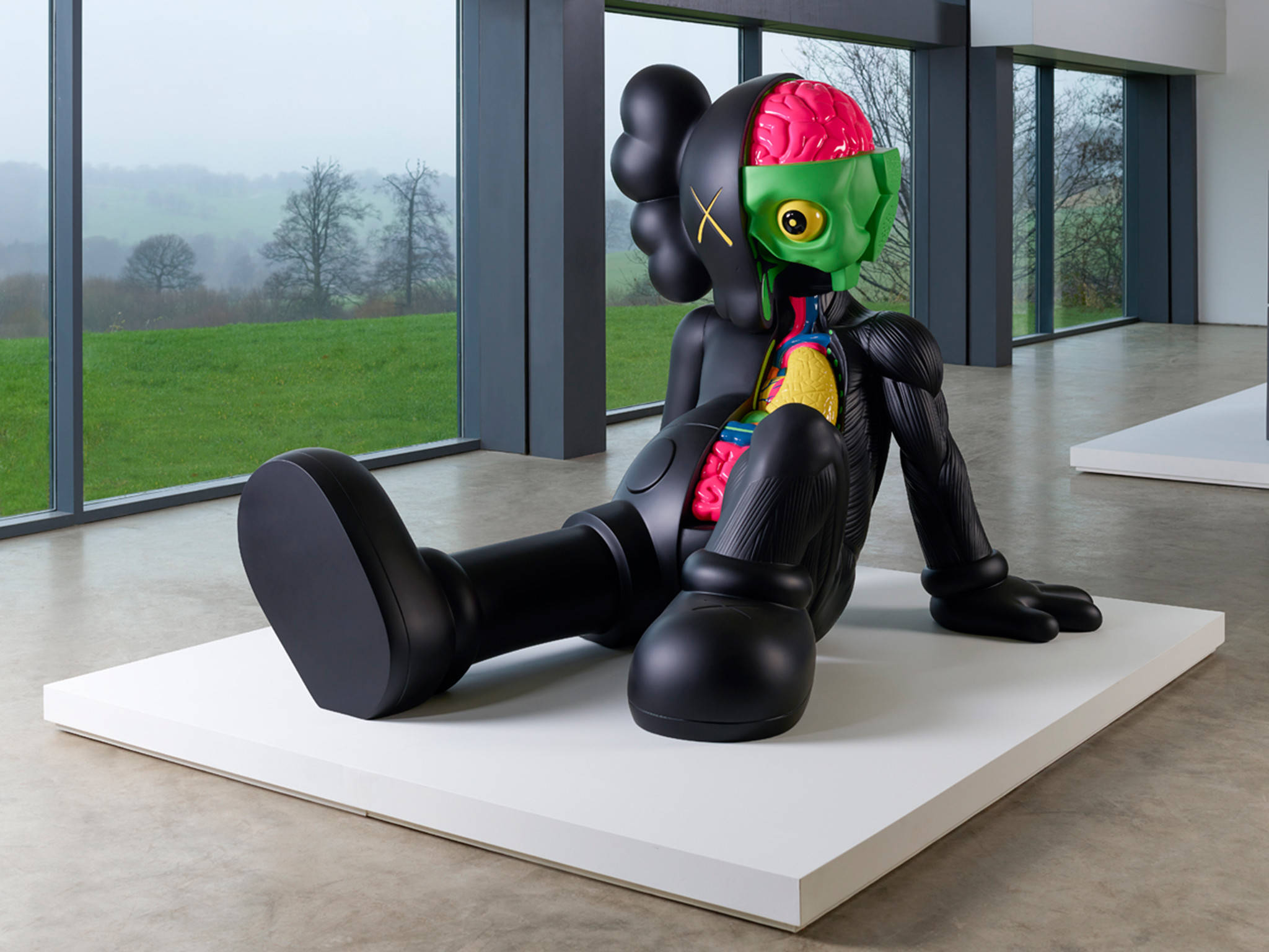 KAWS 桌布 壁紙 — 一個大型黑色卡通風格雕塑，暴露著色彩繽紛的器官，坐在白色底座上，位於現代畫廊內，有大窗戶俯瞰綠色田野。