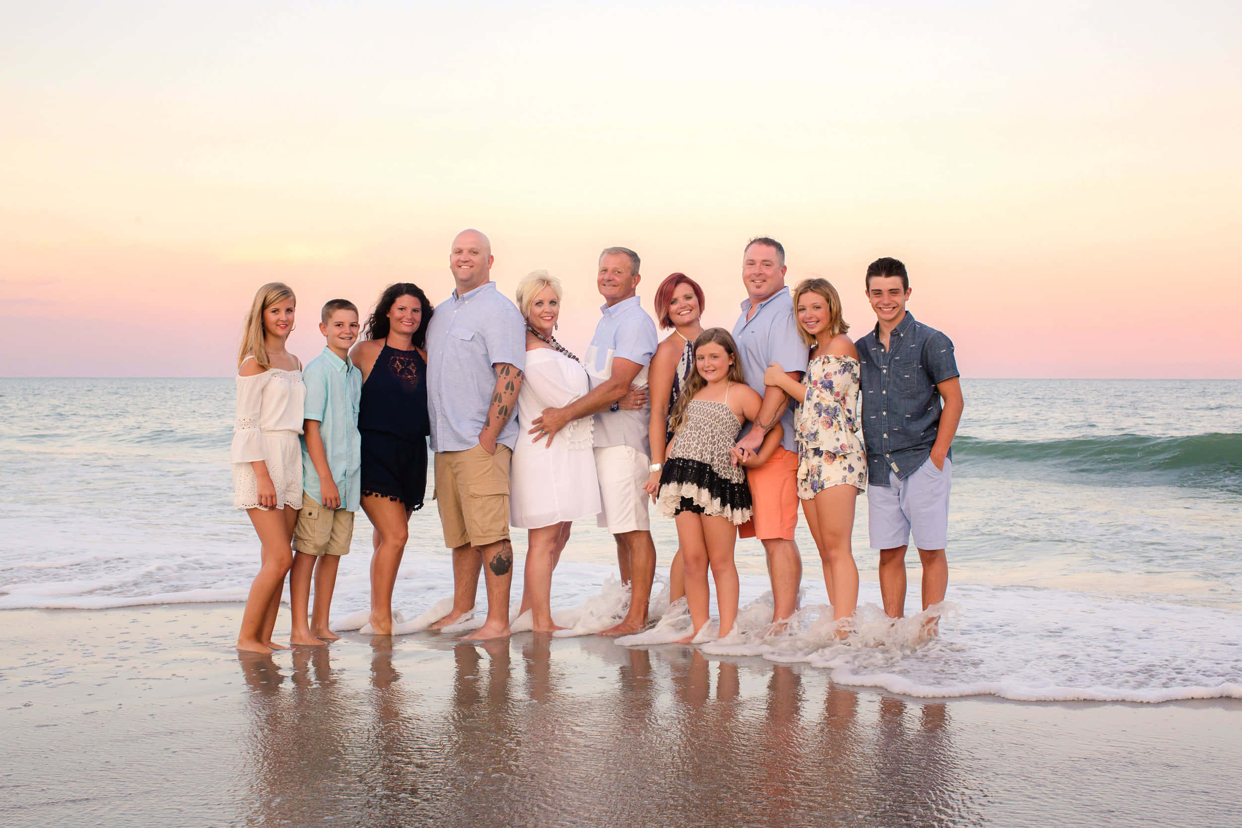 Beach Family 桌布 壁紙 — 一個大家庭在日落時分一起站在濕漉漉的海灘上，溫柔的海浪拍打著他們的腳，身後是柔和的粉橙色天空。