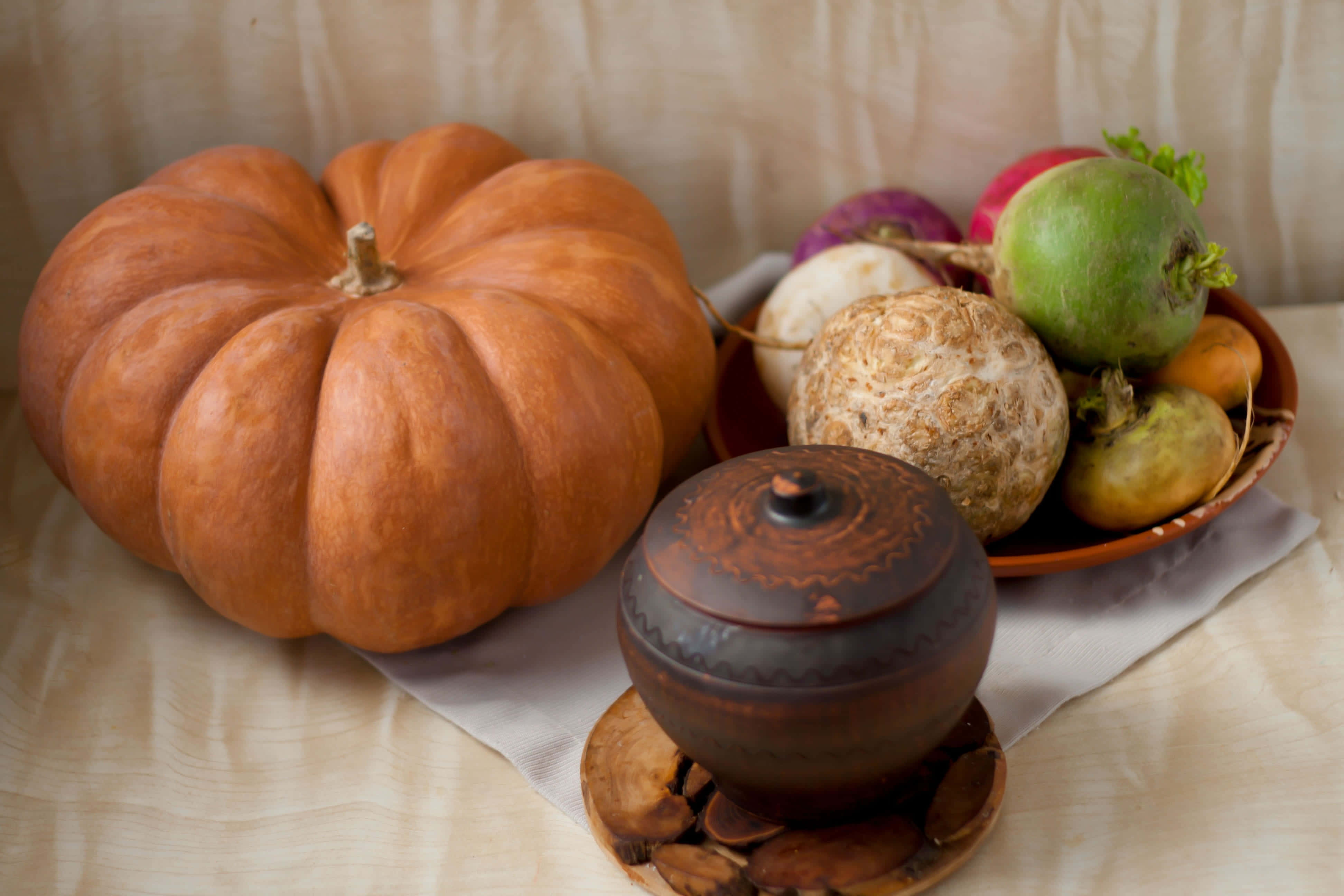 Autumn Food 桌布 壁紙 — 一個大橙色南瓜旁邊放著一碗色彩繽紛的根莖類蔬菜和一個裝飾性陶瓷罐，放在亞麻布上。