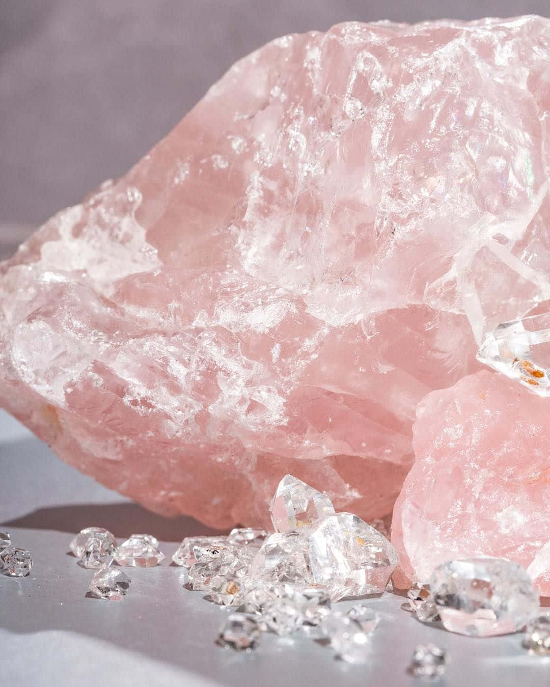 Pink Crystal Quartz 桌布 壁紙 — 一個大的粉紅色玫瑰石英晶體，邊緣參差不齊，散落著透明的寶石，放在光滑的灰色表面上。