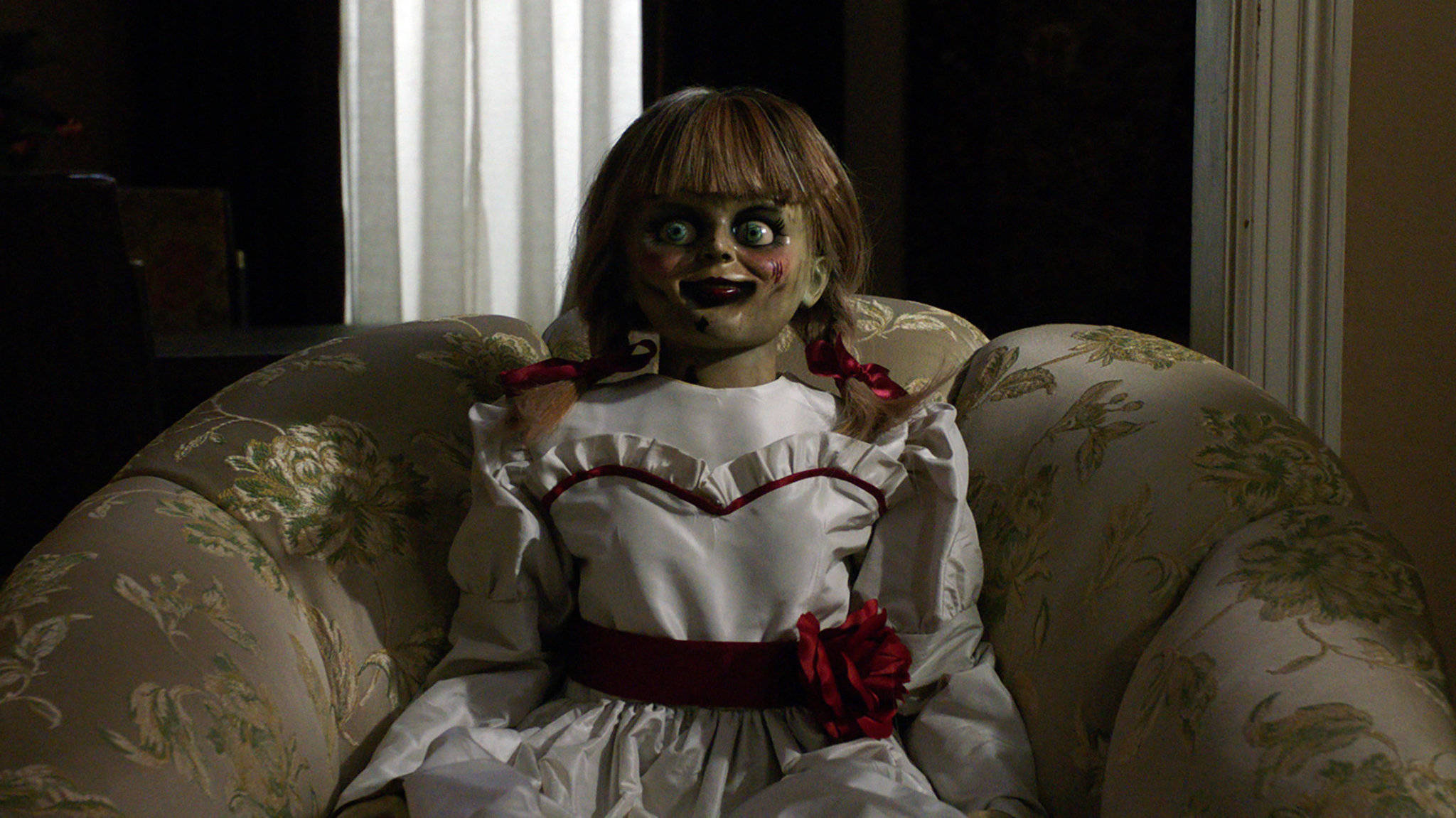 Annabelle 桌布 壁紙 — 一個大眼睛、戴紅色蝴蝶結的恐怖娃娃坐在昏暗房間的花卉圖案沙發上。
