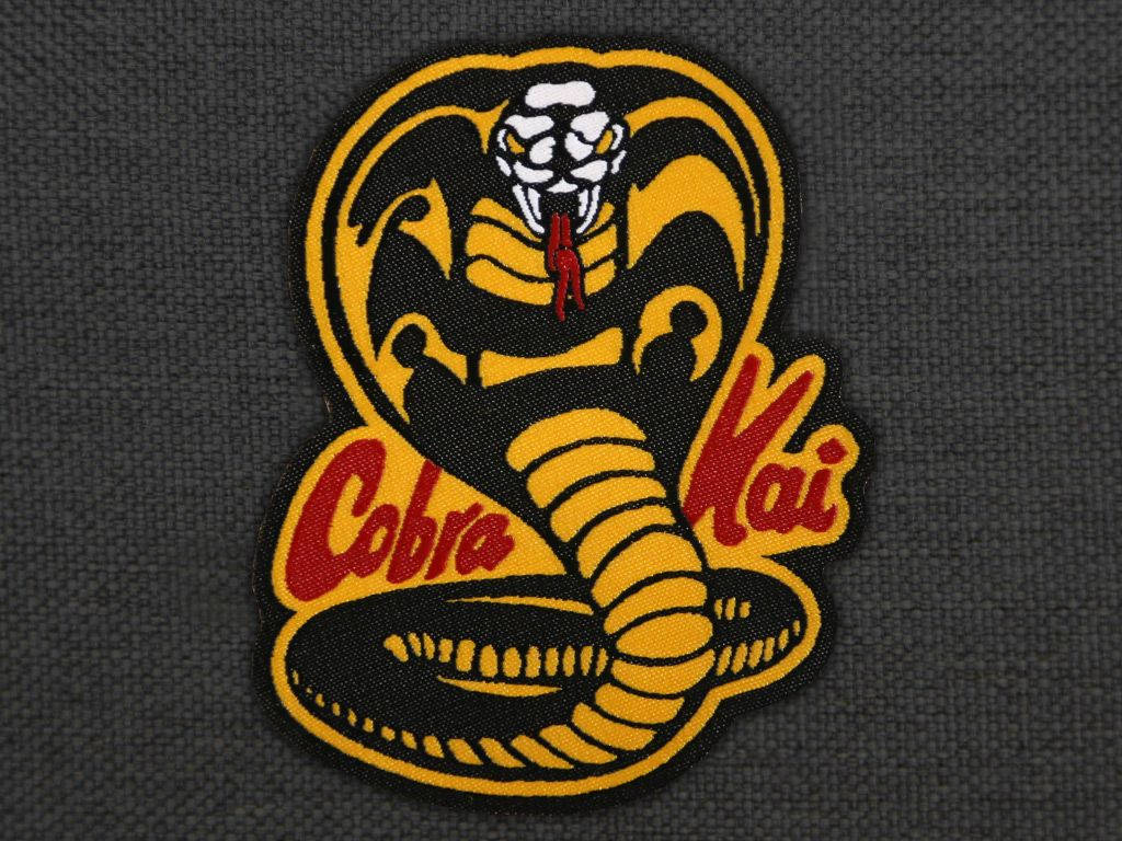 眼鏡蛇道館 桌布 壁紙 — 一個大膽的 Cobra Kai 刺繡徽章，黃黑相間的蛇纏繞在深灰色布料上。