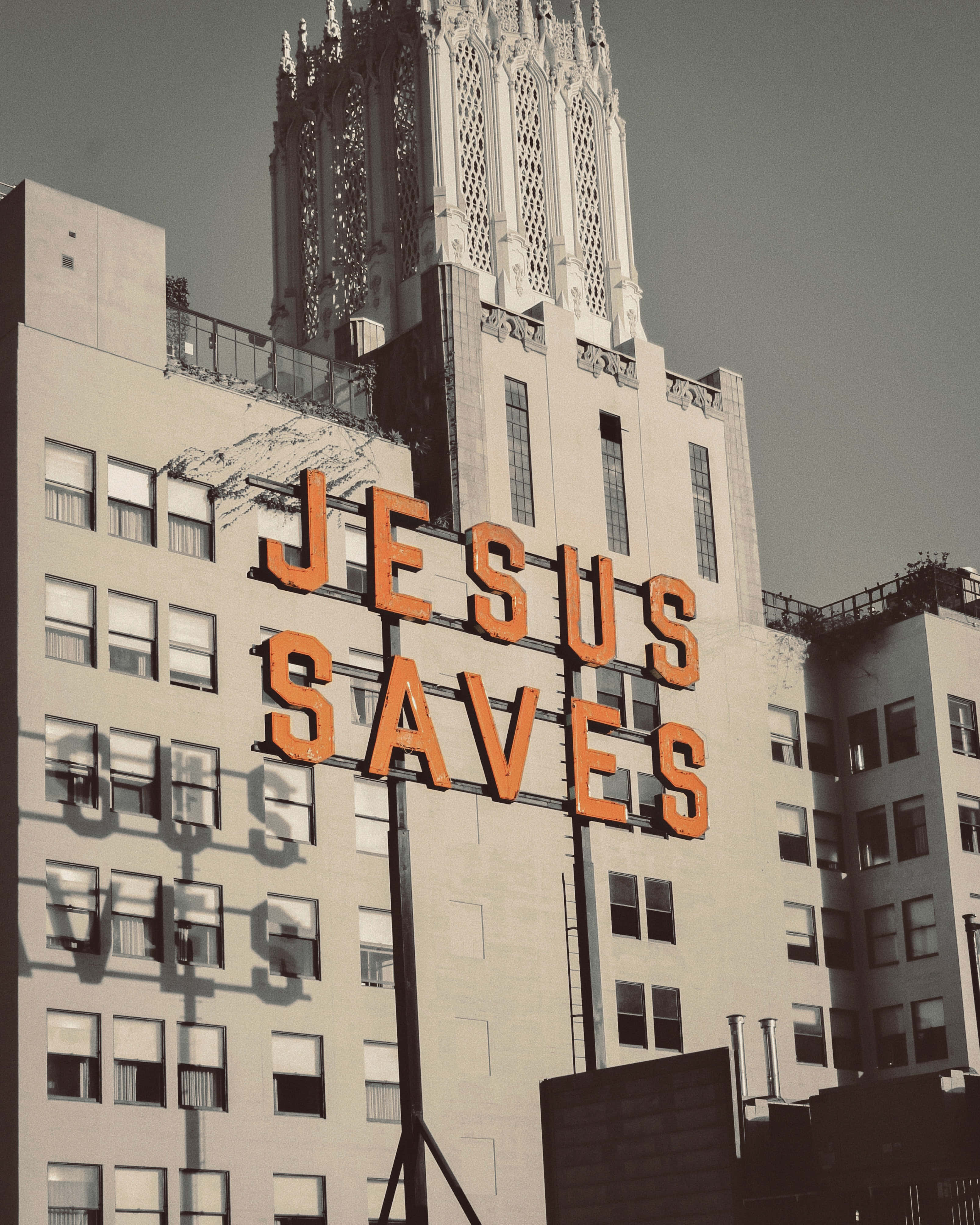 Jesus Saves 桌布 壁紙 — 一個大膽的橙色「JESUS SAVES」標誌在樸素的城市建築和哥德式塔樓襯托下，在陰沉天空下格外突出。
