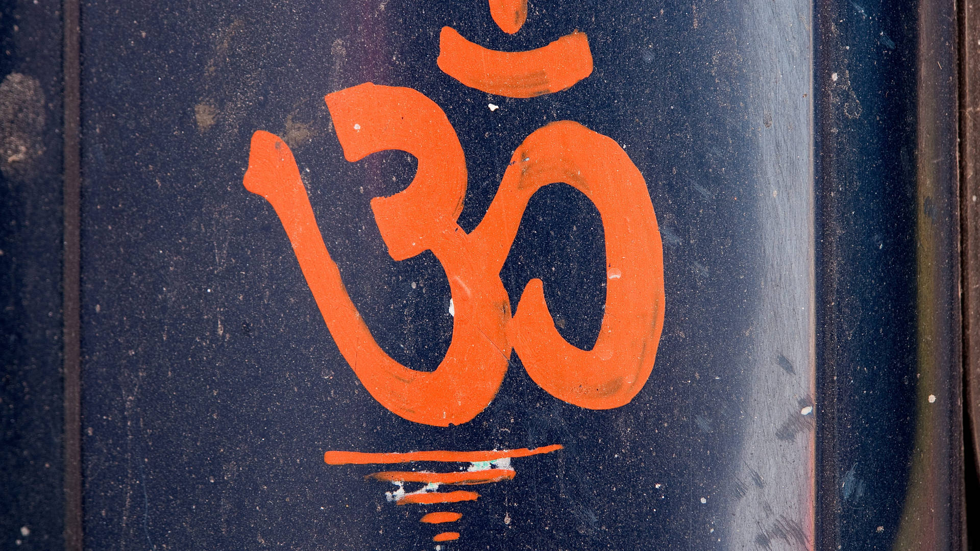 Om 桌布 壁紙 — 一個大膽的橙色 Om 符號，繪製在風化的深藍色金屬表面上，帶有可見的紋理和磨損。