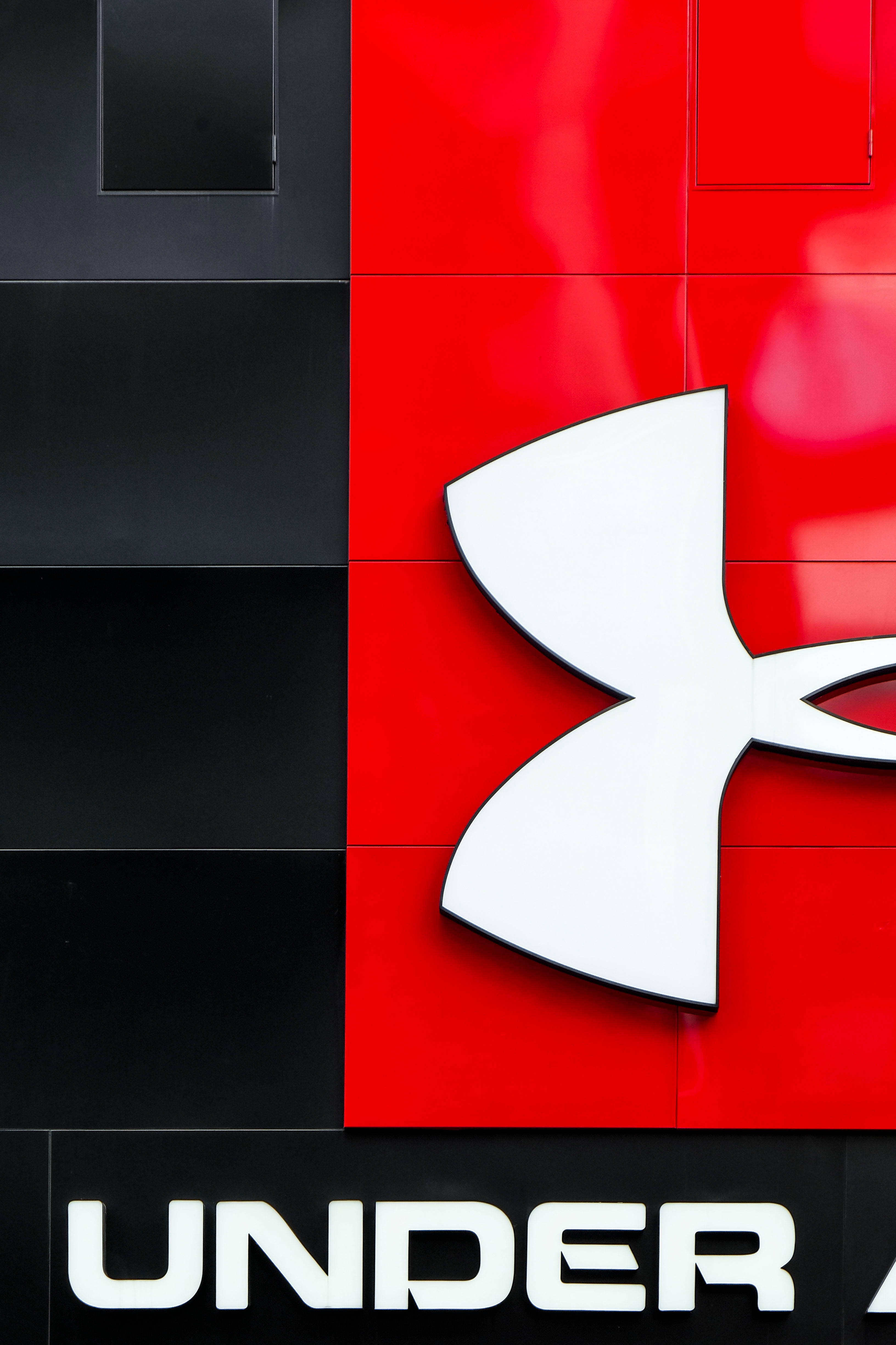 Under Armour 桌布 壁紙 — 一個大膽的白色 Under Armour 標誌在充滿活力的紅色和時尚的黑色瓷磚牆上脫穎而出。