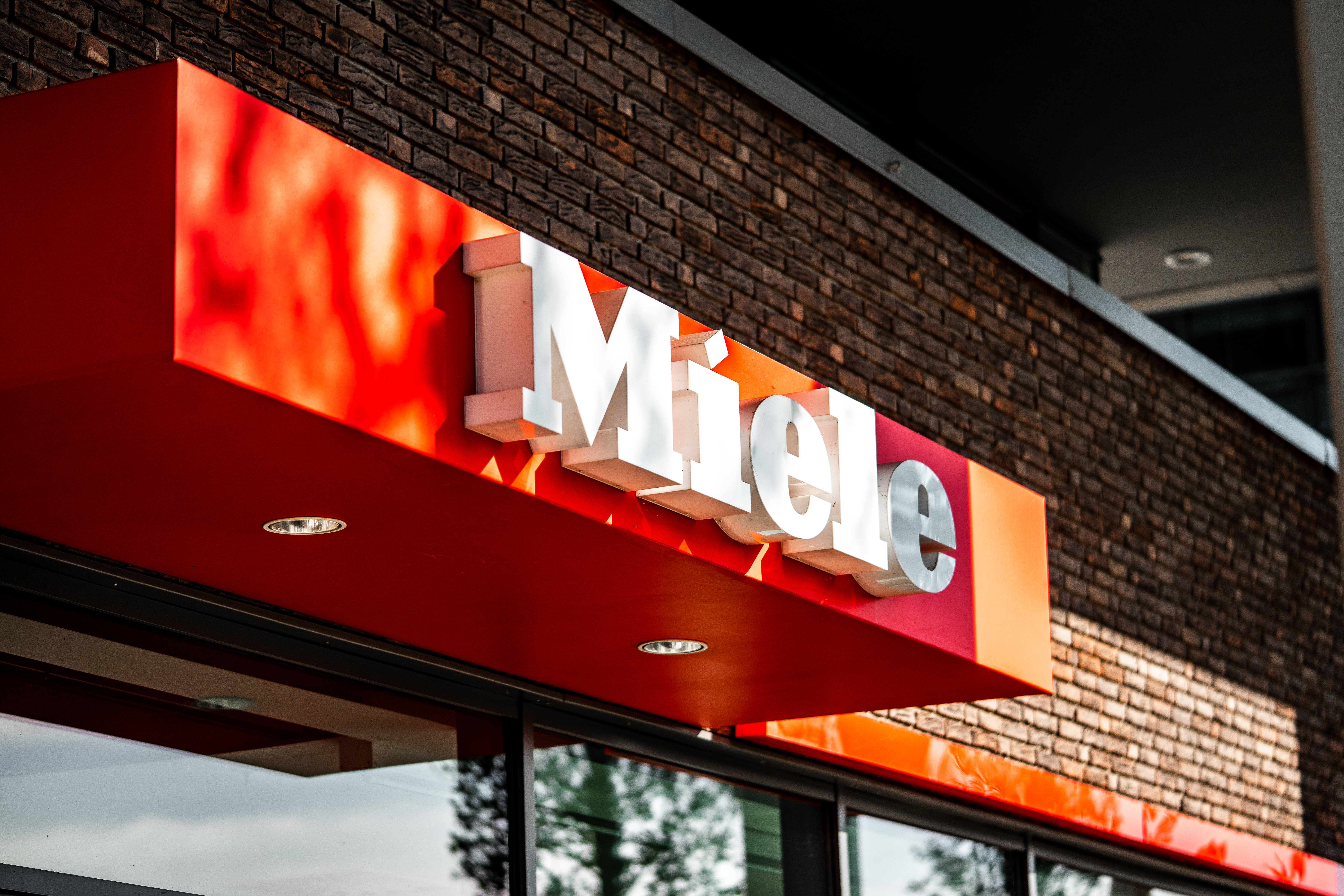標誌 桌布 壁紙 — 一個大膽的紅色 Miele 標誌，帶有白色字體，安裝在磚砌建築外牆上，在明亮的日光下。
