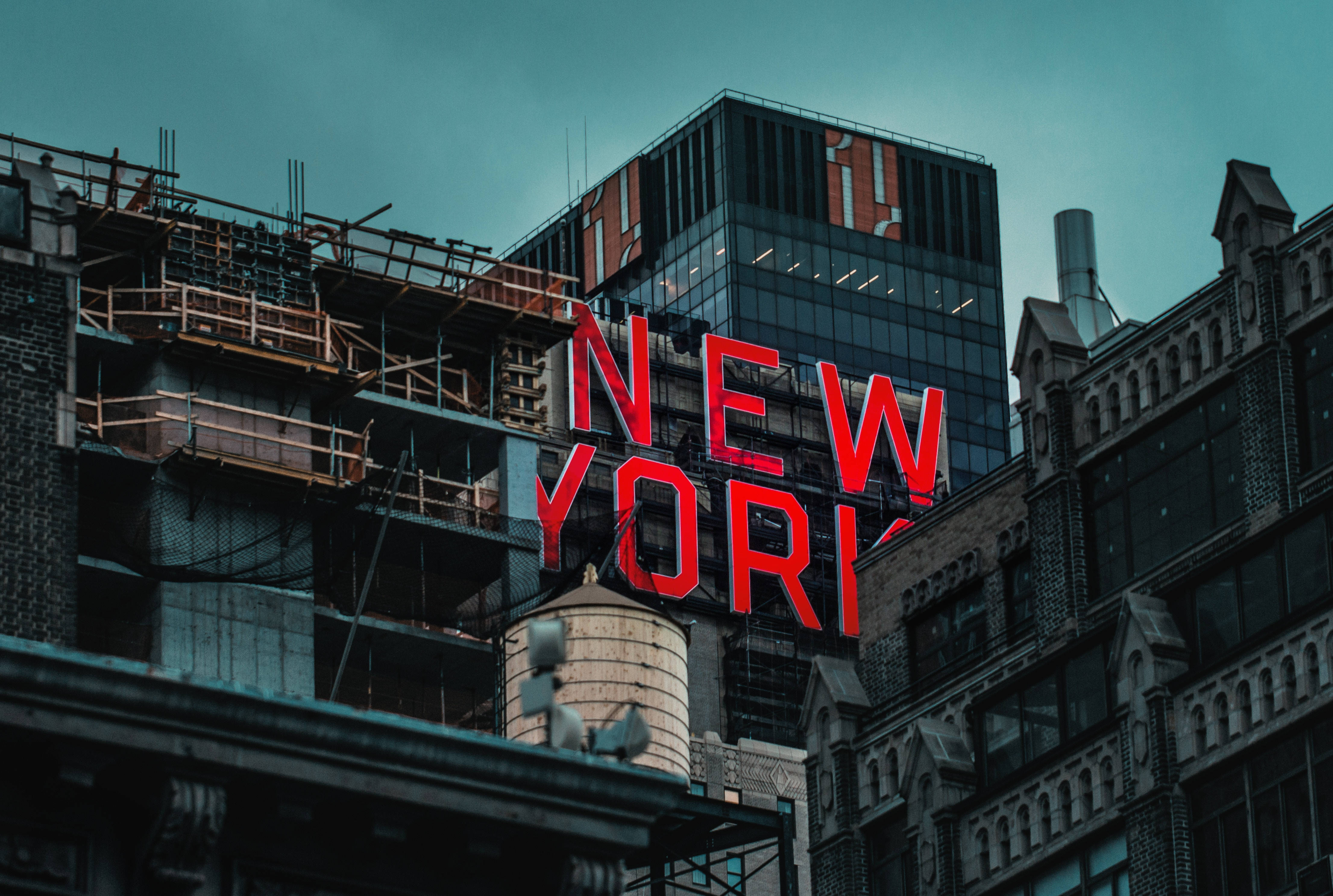 New 桌布 壁紙 — 一個大膽的紅色「NEW YORK」標誌在憂鬱的藍綠色天空下發光，下方是歷史磚砌建築和現代玻璃塔樓的混合體，正在建設中。