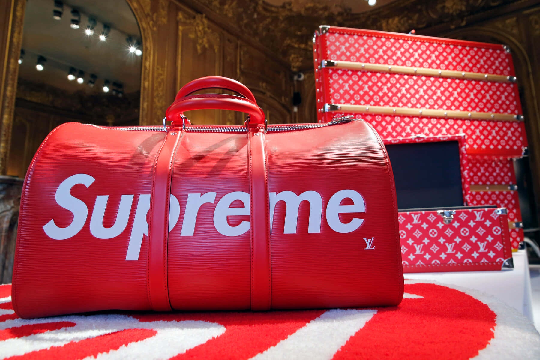 supreme 桌布 壁紙 — 一個大膽的紅色 Supreme x Louis Vuitton 行李袋放在相配的地毯中央，兩側是堆疊的紅色 monogram 行李箱，在一個華麗的金碧輝煌的房間裡。