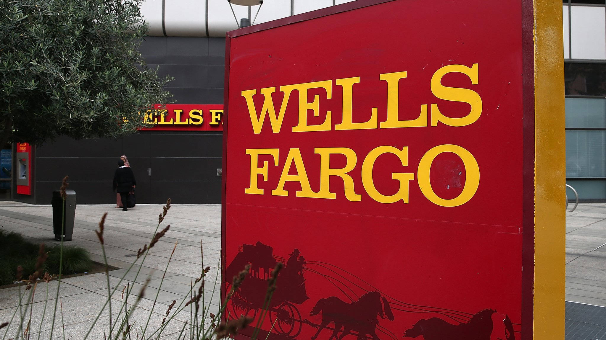 標誌 桌布 壁紙 — 一個大膽的紅色 Wells Fargo 標誌，帶有黃色字體和馬車剪影，矗立在一棟現代建築前，有人走過。