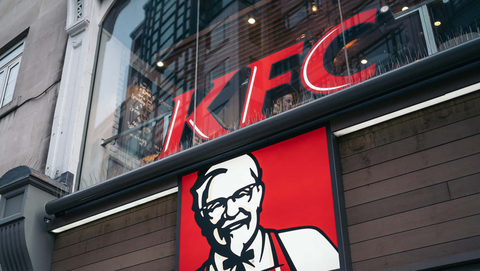肯德基 桌布 壁紙 — 一個大膽的紅色KFC招牌，上面有桑德斯上校標誌性的笑臉，點綴著現代都市餐廳的外牆。
