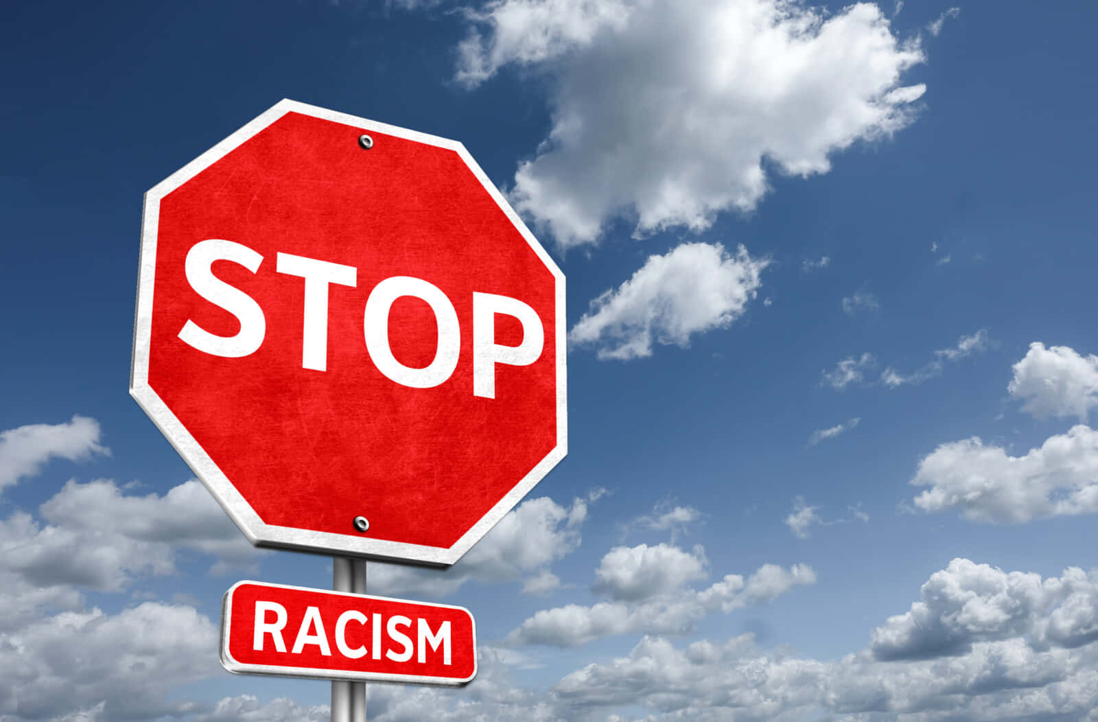 stop 桌布 壁紙 — 一個大膽的紅色停車標誌，上面有「STOP」，下面有一個較小的「RACISM」標誌，以晴朗的藍天和蓬鬆的白雲為背景。