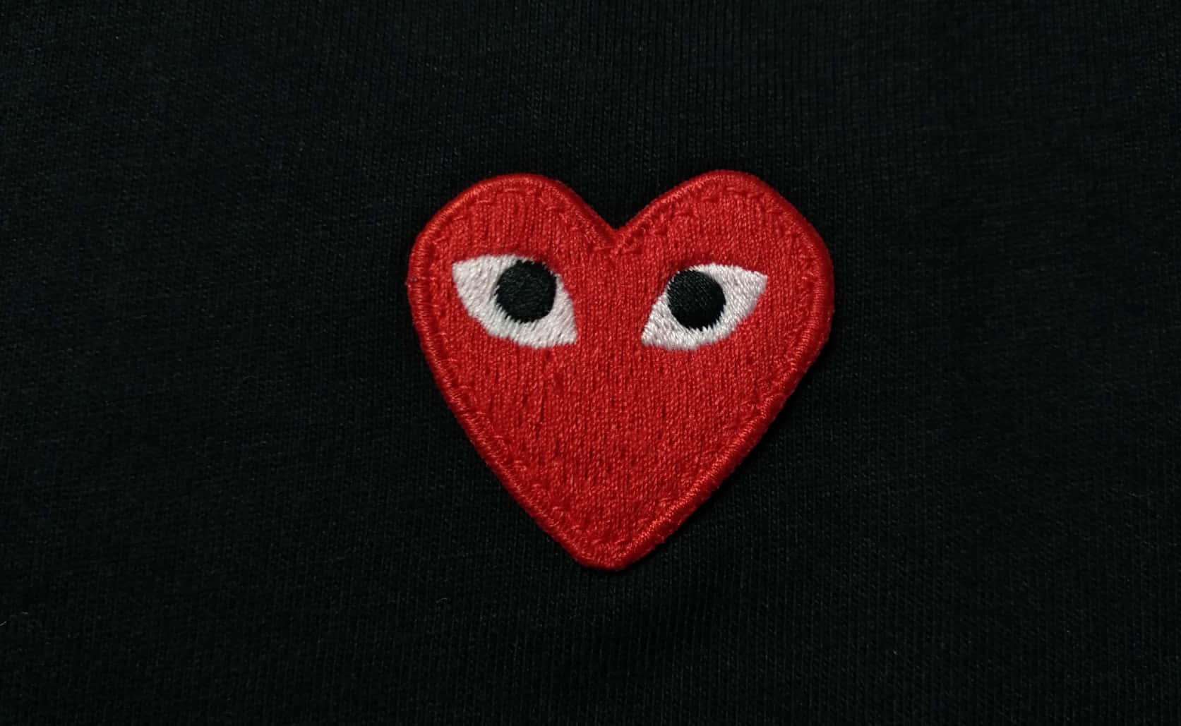 愛心 桌布 壁紙 — 一個大膽的紅色刺繡心形補丁，帶有白色眼睛，縫在黑色布料背景上。