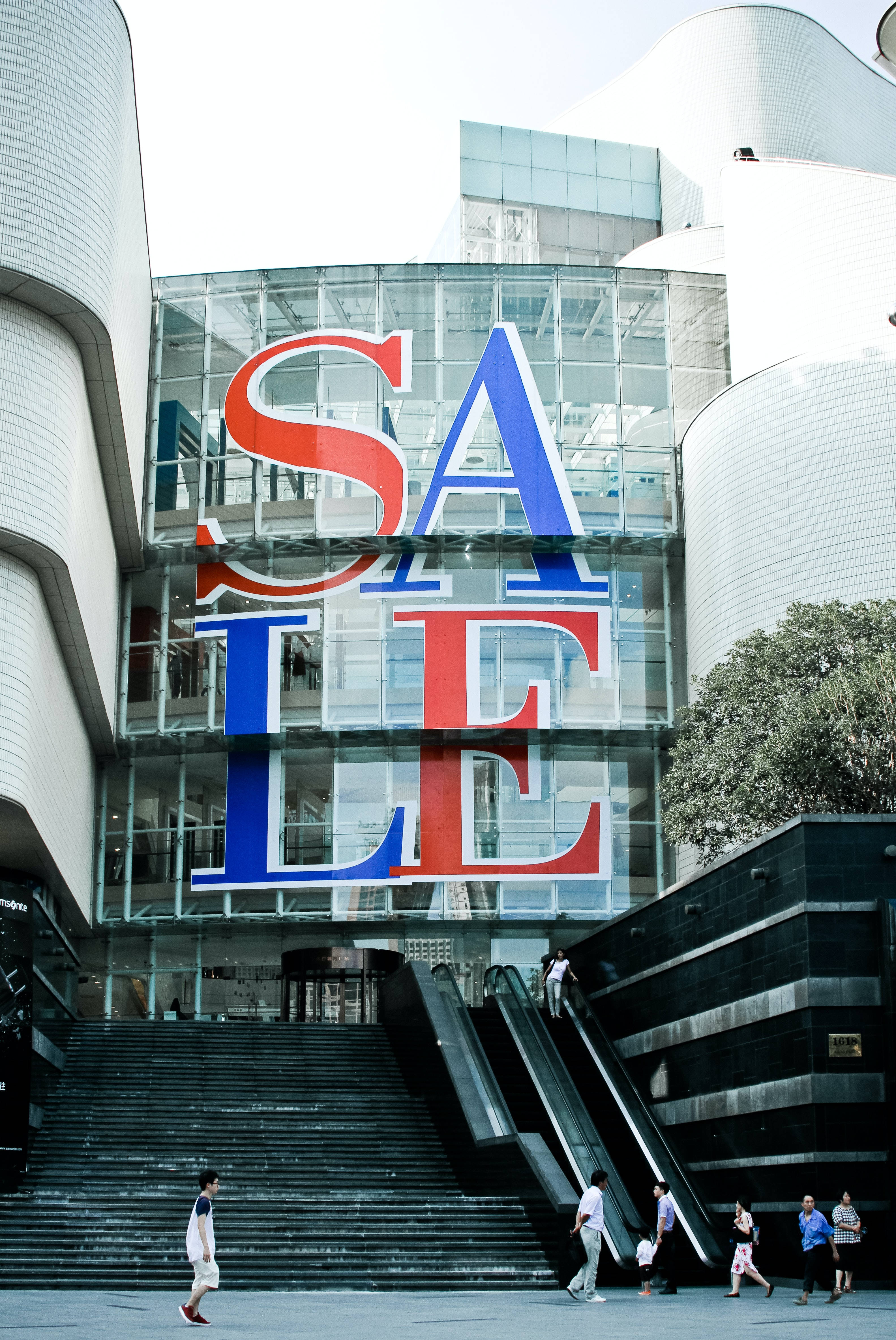 Sales 桌布 壁紙 — 一個大膽的紅色和藍色SALE標誌主導著一座現代玻璃建築，帶有彎曲的白色建築和附近行走的人們。