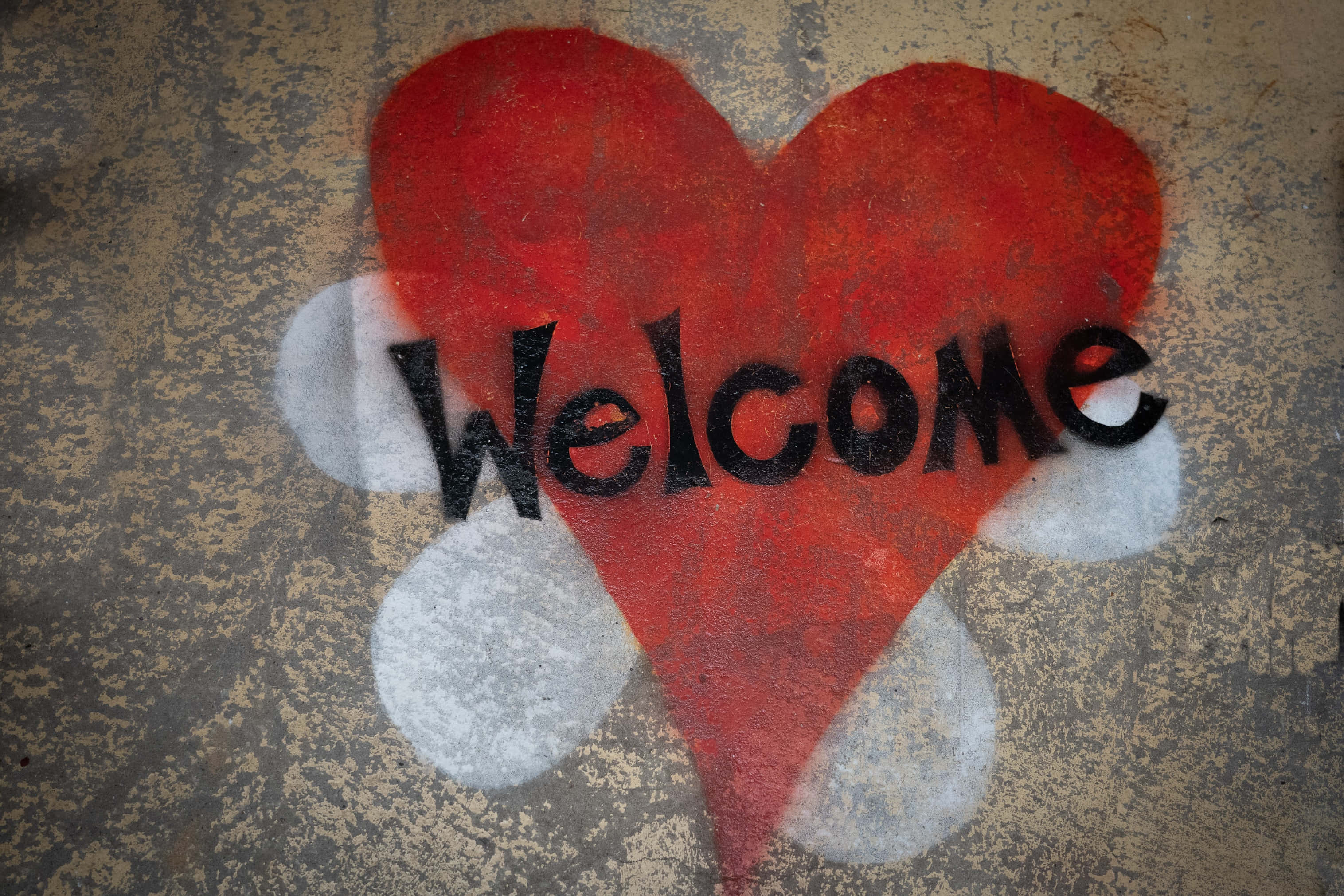 Welcome 桌布 壁紙 — 一個大膽的紅色心形，上面用黑色噴漆寫著「Welcome」，背景是帶有白色圓形裝飾的風化混凝土牆。