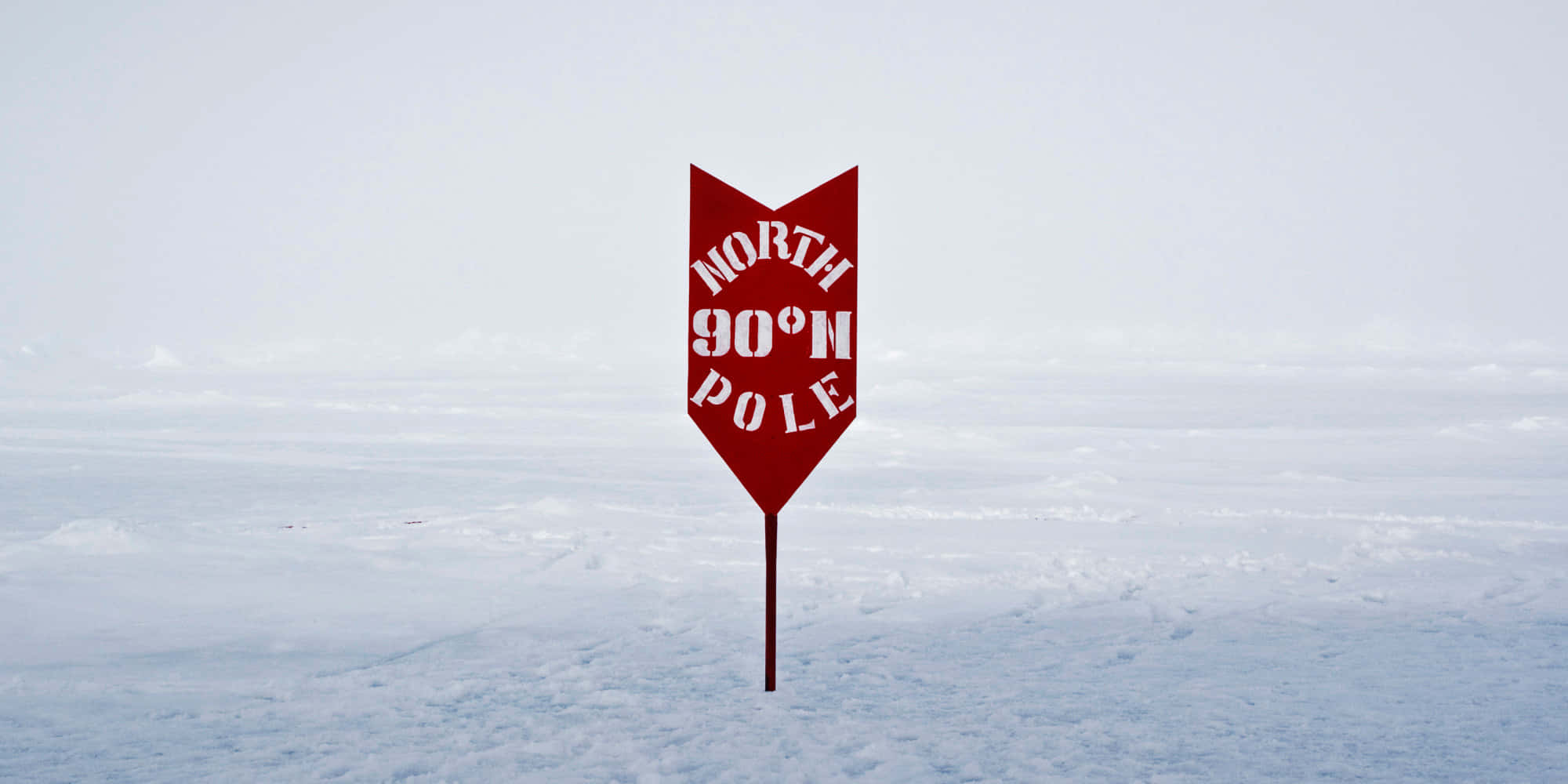 North Pole 桌布 壁紙 — 一個大膽的紅色箭頭形標誌，上面寫著「NORTH 90°N POLE」，獨自矗立在廣闊的雪白北極景觀中，天空蒼白。