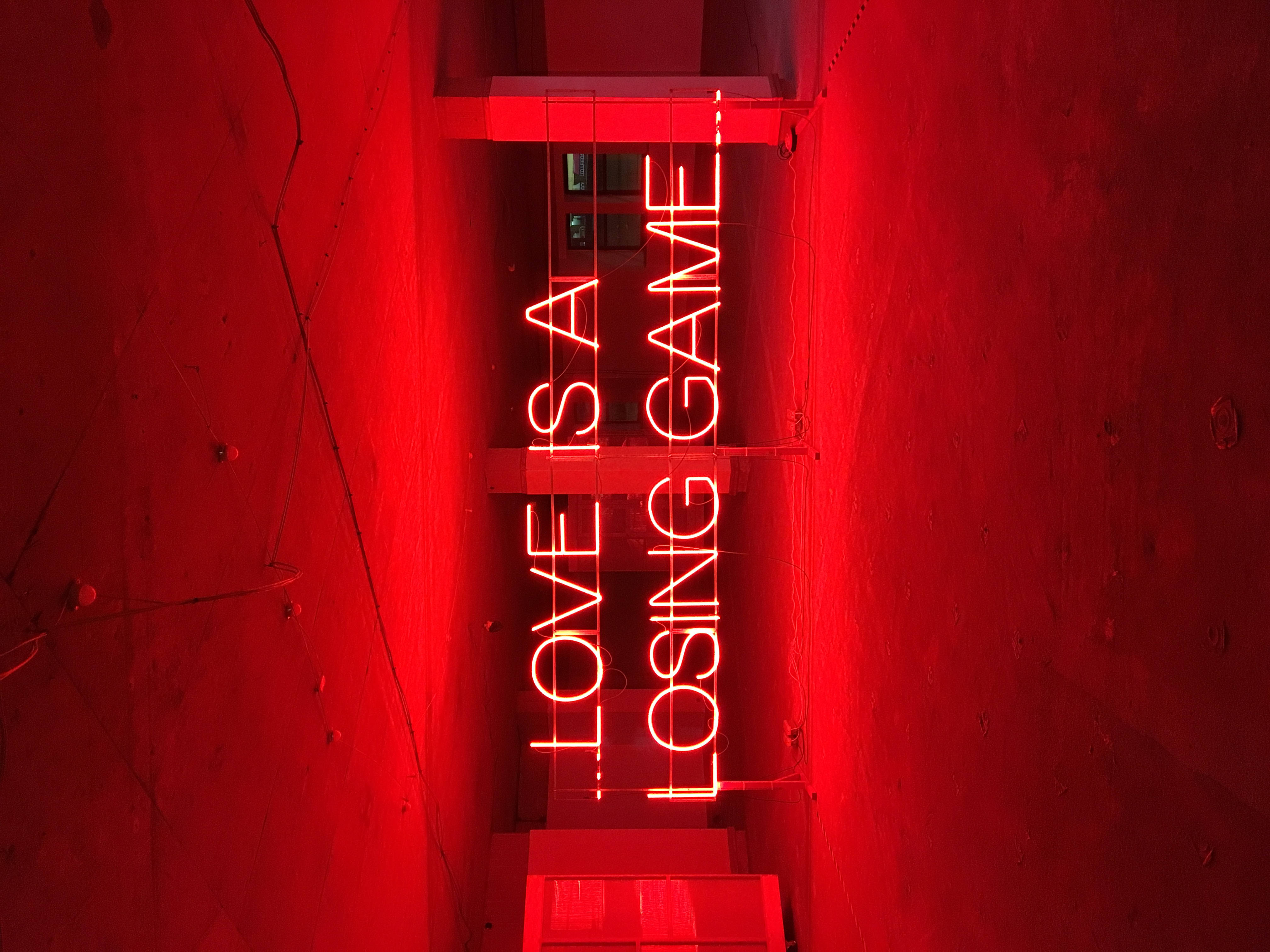 紅色動圖 桌布 壁紙 — 一個大膽的紅色霓虹燈牌，上面寫著「LOVE IS A LOSING GAME」，在光線昏暗的城市小巷中，映照在紋理混凝土牆上。