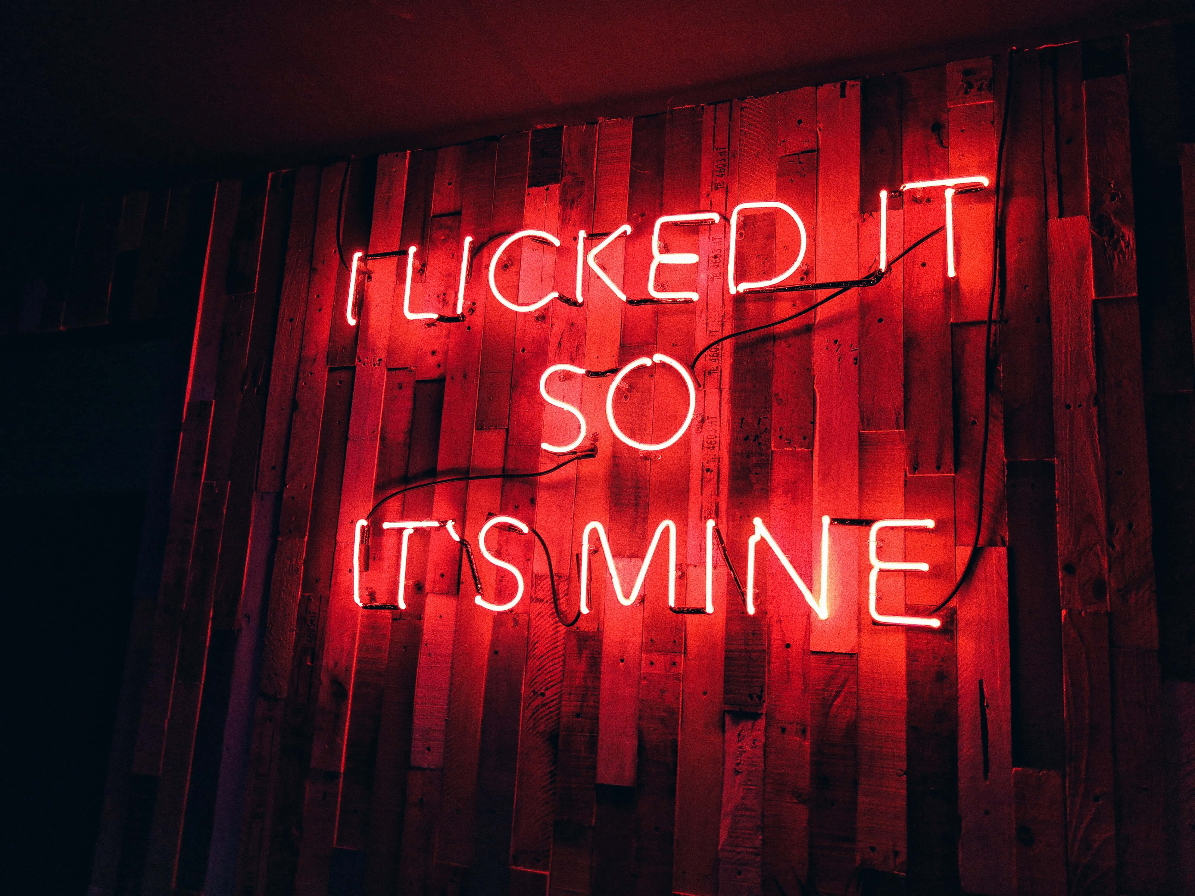 Red Neon 桌布 壁紙 — 一個大膽的紅色霓虹燈牌，上面寫著 'I LICKED IT SO IT'S MINE'，在昏暗的空間中對著質樸的木牆發光。