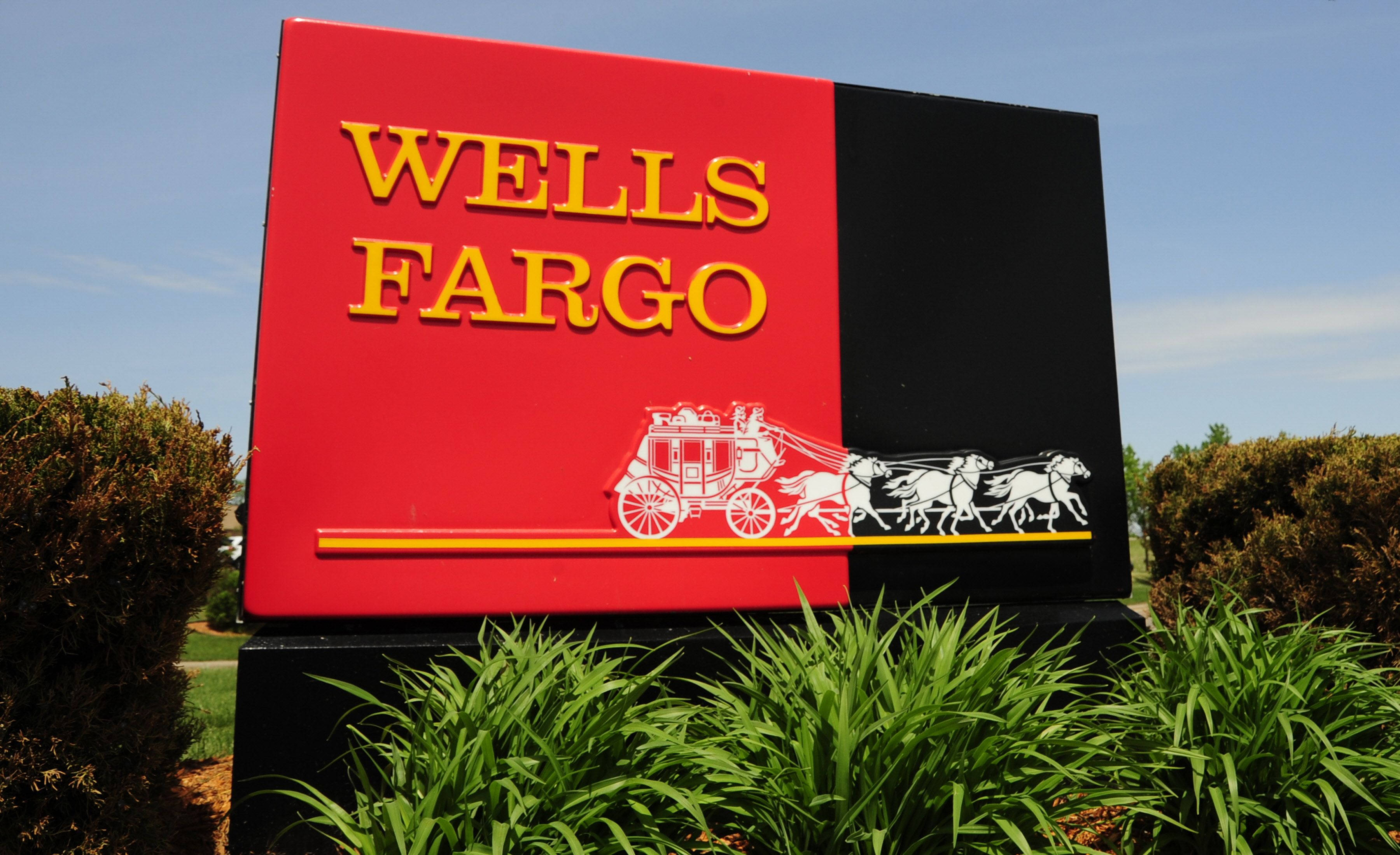 標誌 桌布 壁紙 — 一個大膽的紅黑 Wells Fargo 標誌，帶有白色驛馬車標誌，在晴朗的藍天下高高聳立，周圍環繞著茂密的綠色灌木。