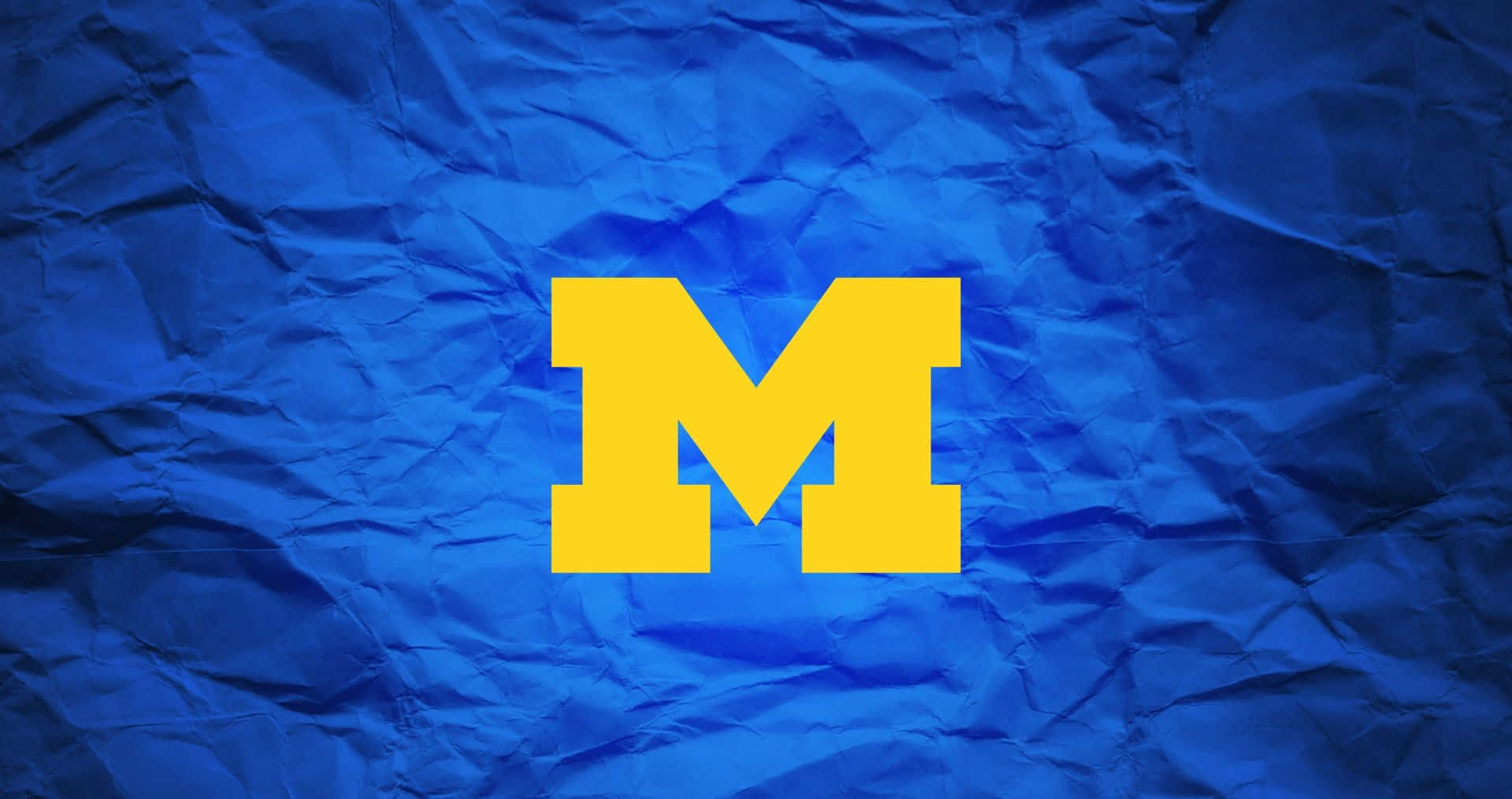 Michigan Wolverines 桌布 壁紙 — 一個大膽的黃色塊狀M標誌，居中放置在帶有紋理的皺巴巴的皇家藍色紙上。