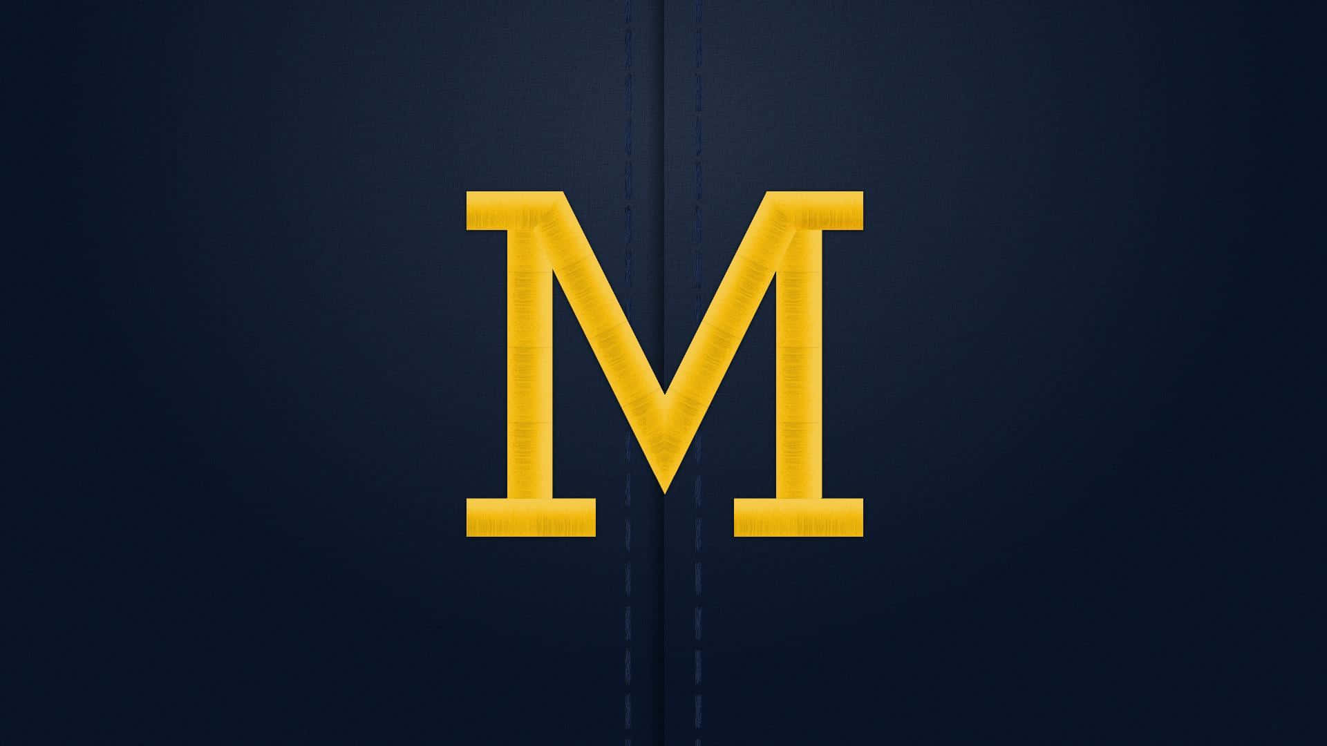 Michigan Wolverines 桌布 壁紙 — 一個大膽的黃色字母M縫在深藍色布料背景上，縫線清晰可見。