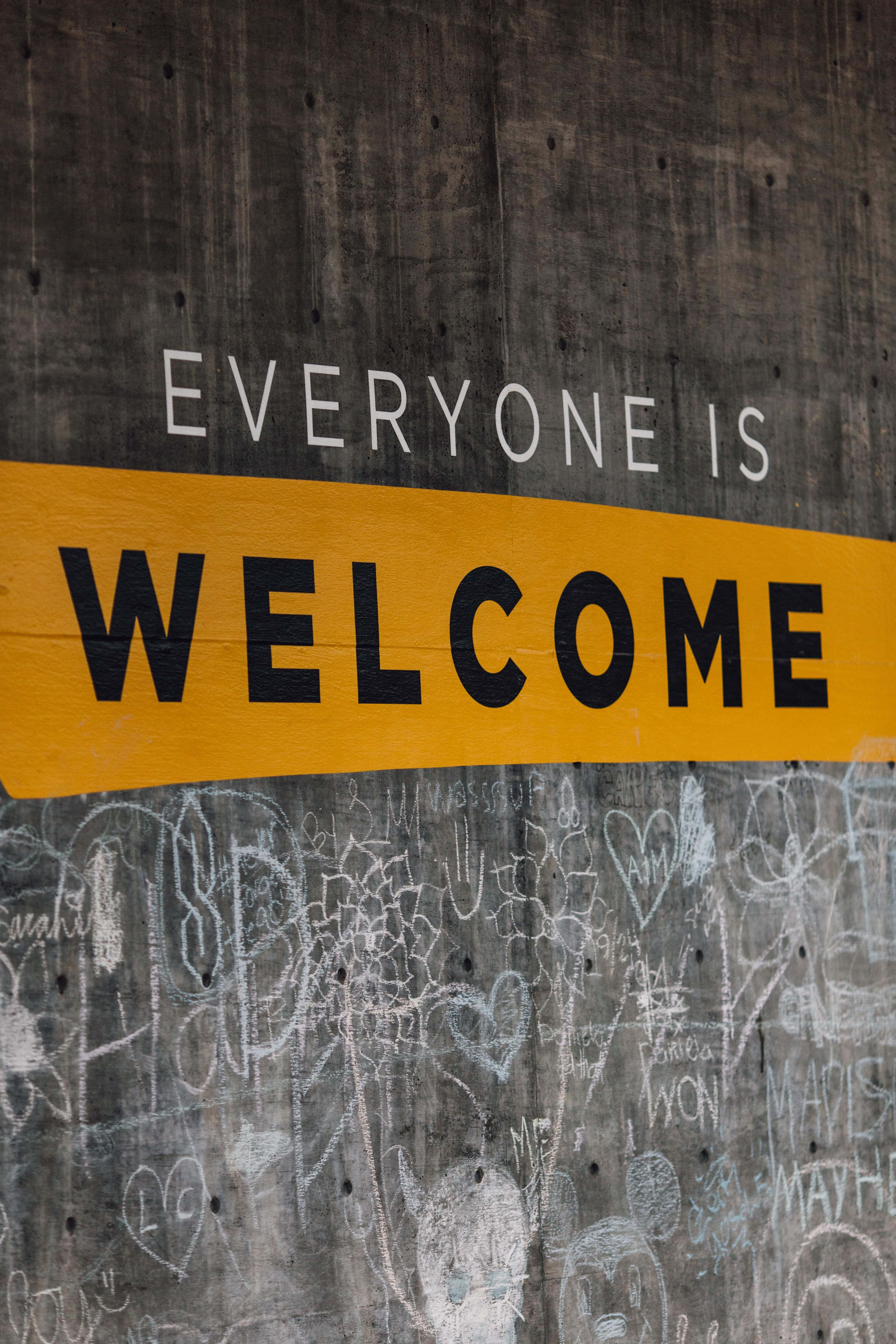 Welcome 桌布 壁紙 — 一個大膽的黃色標誌寫著 everyone is welcome，與佈滿粉筆塗鴉的風化混凝土牆形成對比。