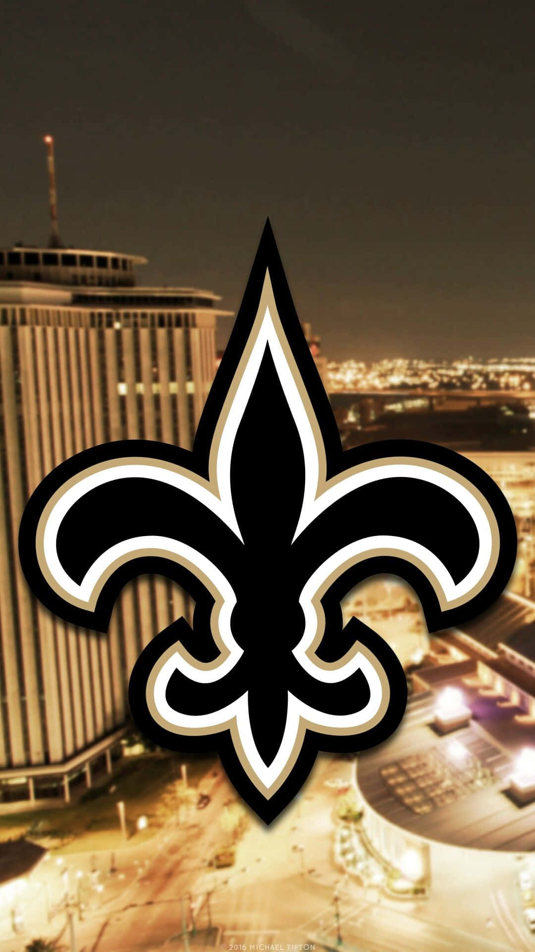 NFL Saints 桌布 壁紙 — 一個大膽的黑金色百合花標誌主導著夜晚的城市景觀，建築物和街道燈火通明。