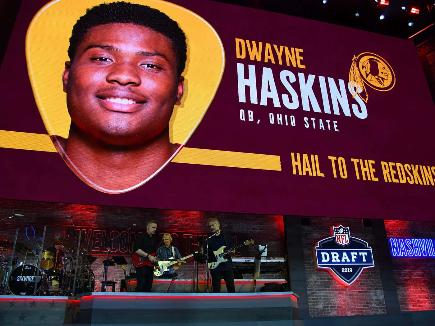 Dwayne Haskins 桌布 壁紙 — 一個大螢幕顯示Dwayne Haskins在吉他形狀的框架中微笑，位於2019年NFL選秀舞台上，下方有音樂家表演。