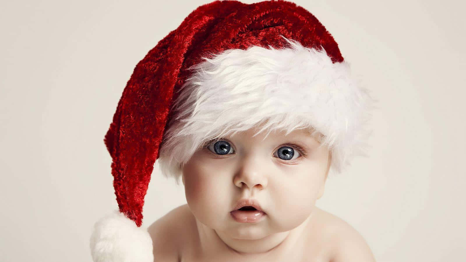 cute baby 桌布 壁紙 — 一個天真無邪的嬰兒戴著紅色天鵝絨聖誕帽，帽緣有白色毛邊，背景是柔和的米色。
