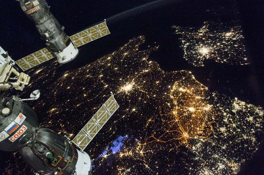 Satellite 桌布 壁紙 — 一個太空站懸浮在地球發光的夜間城市上方，複雜的光線圖案照亮了下方的各大洲。