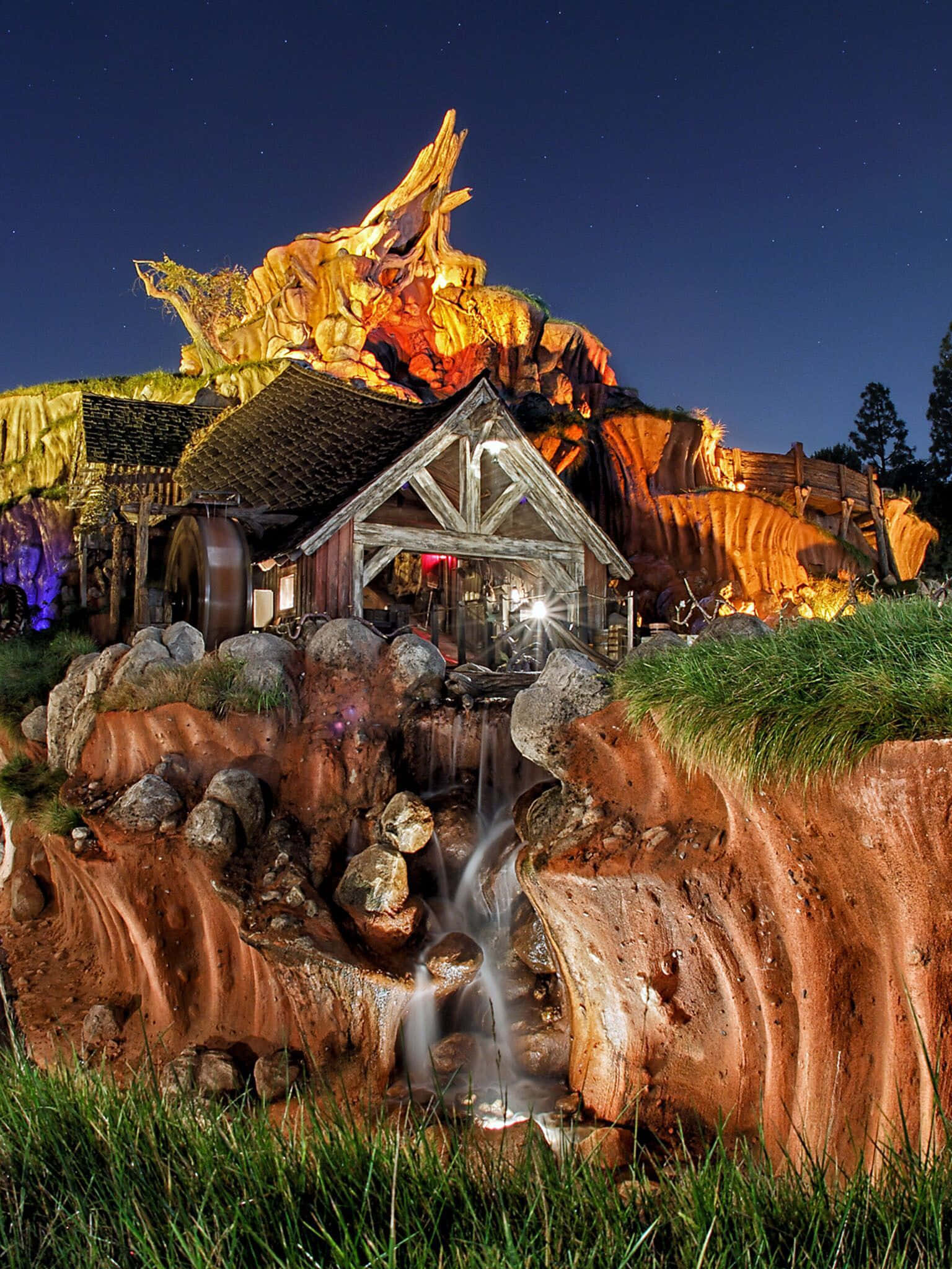 Splash Mountain 桌布 壁紙 — 一個奇特的夜景場景，展示了一座質樸的木製磨坊，坐落在岩石峽谷中，周圍是發光的橙色懸崖和星空下的瀑布。