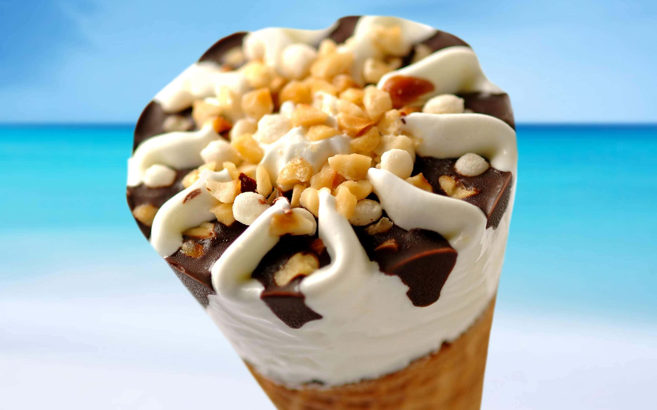 Ice Cream 桌布 壁紙 — 一個奢華的冰淇淋甜筒，上面有巧克力漩渦、碎堅果和鮮奶油，背景是明亮的沙灘。