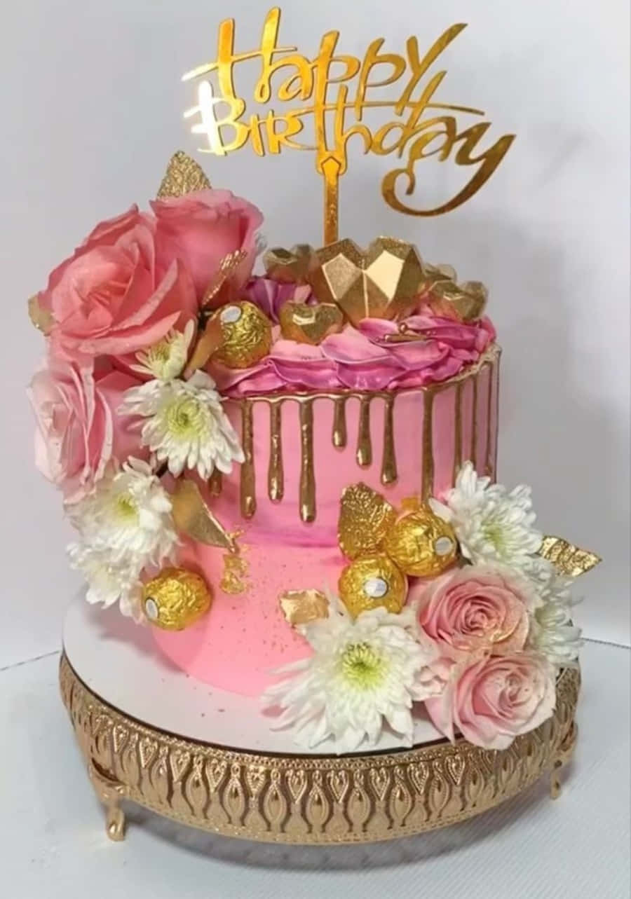 Happy Birthday Cake 桌布 壁紙 — 一個奢華的粉紅色生日蛋糕，飾有金色滴紋、花朵和費列羅巧克力，頂部放有金色「生日快樂」裝飾。