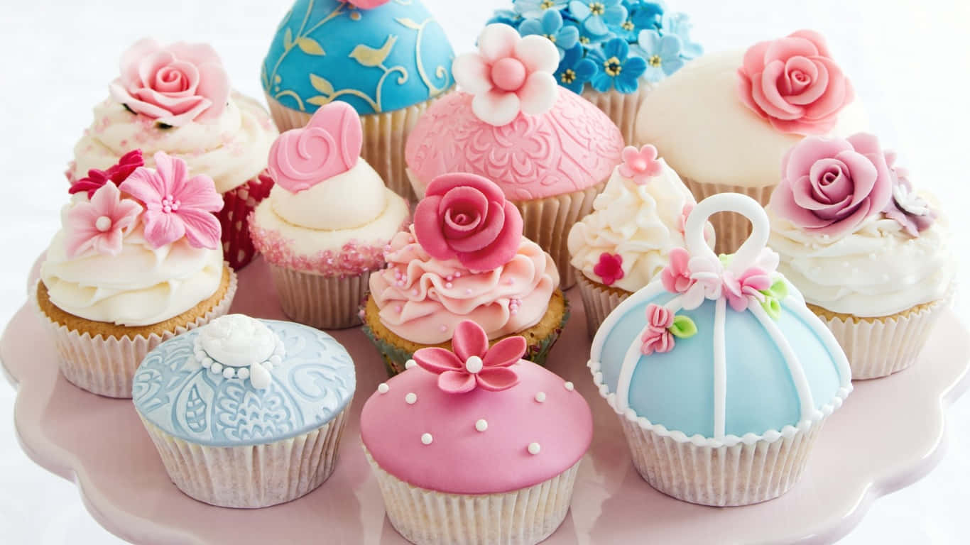 1366x768 Pastries 桌布 壁紙 — 一個奢華的組合，包含精心裝飾的杯子蛋糕，點綴著粉紅玫瑰、藍色翻糖和精緻的糖花，擺放在粉彩粉紅色的盤子上。