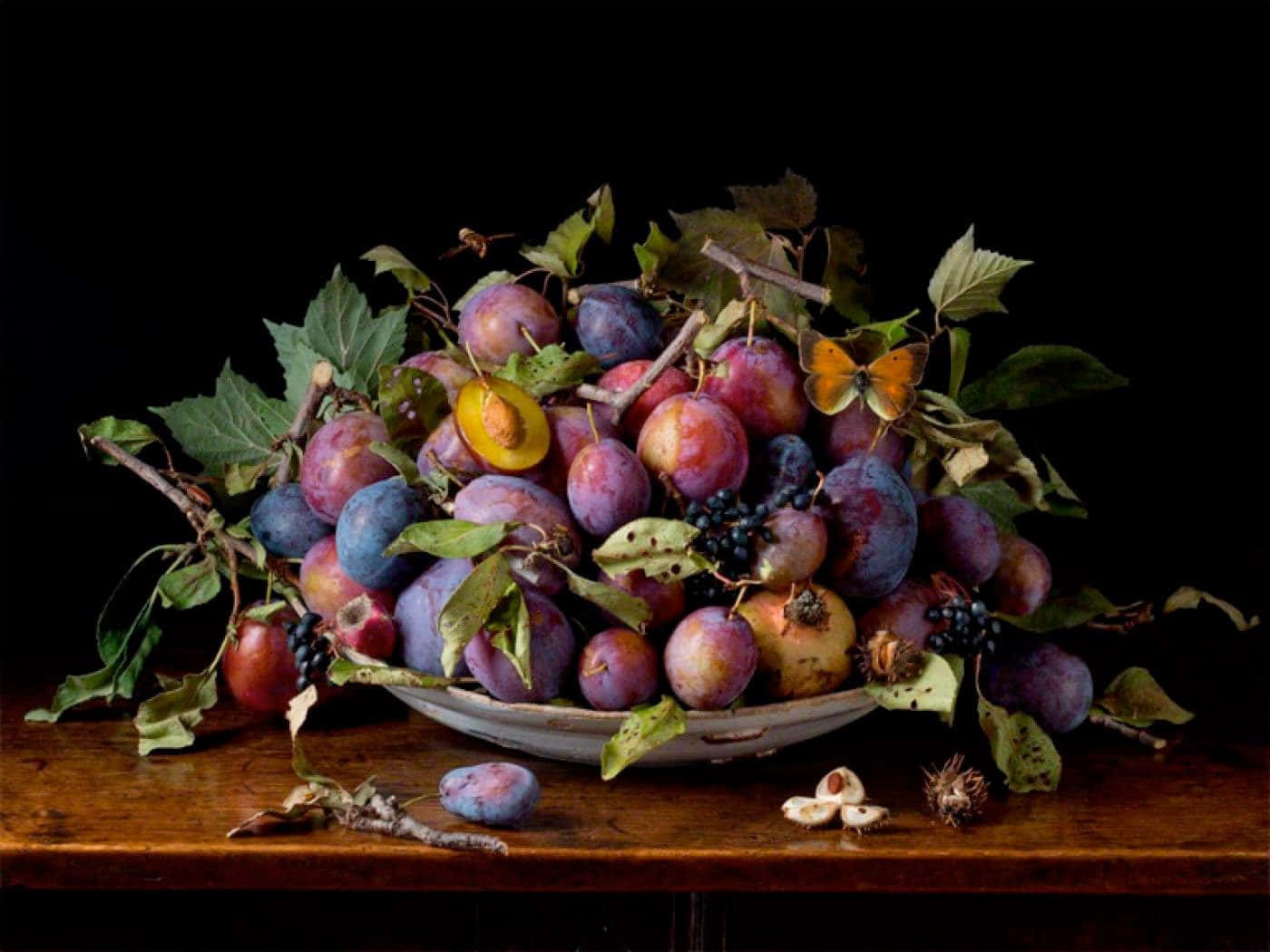 Fruits 桌布 壁紙 — 一個奢華的靜物畫，碗裡裝滿李子、葡萄和葉子，一隻蝴蝶停在頂部，背景為黑色。