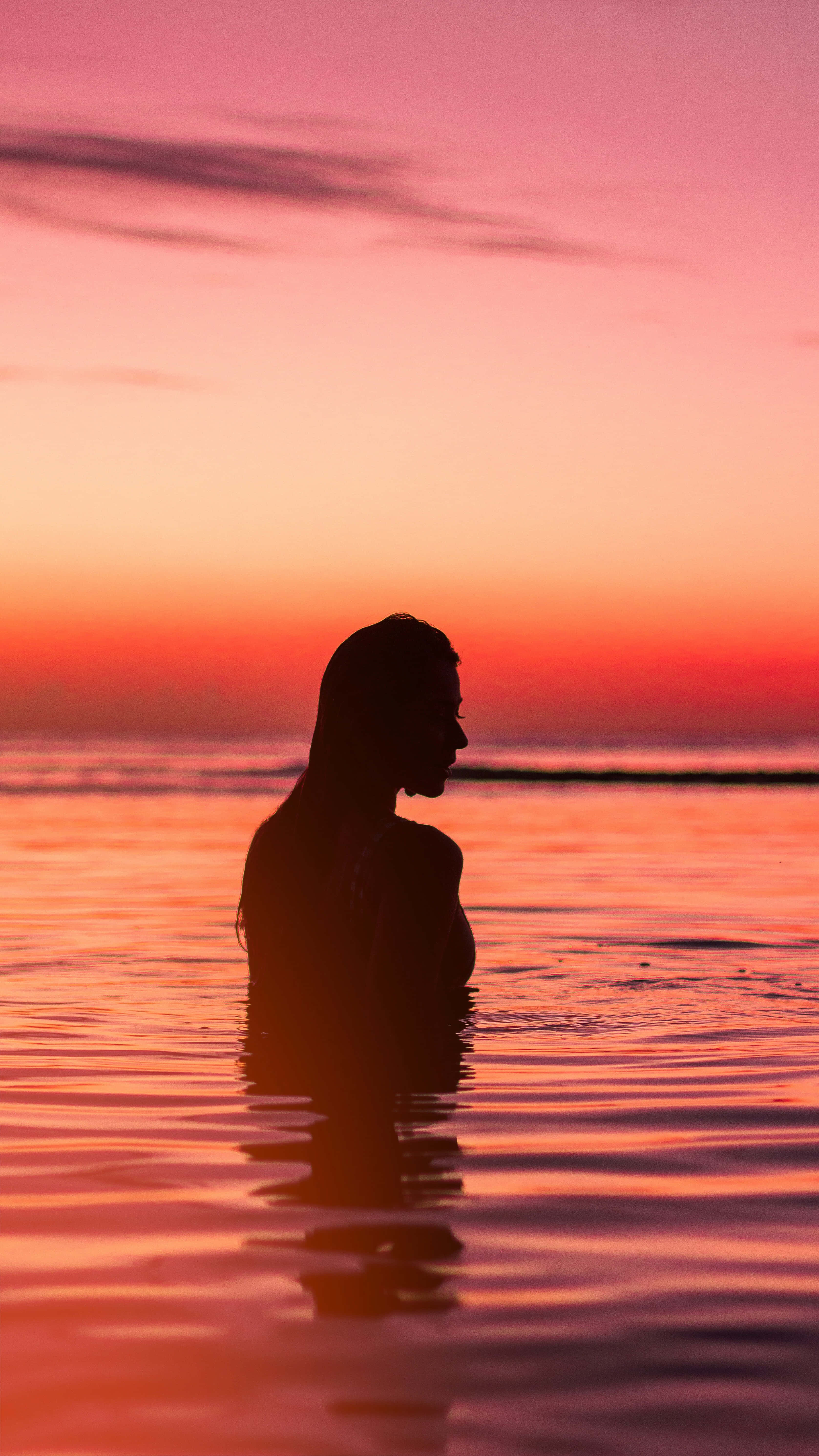 Silhouette 桌布 壁紙 — 一個女人在平靜的海水中站立，背景是帶有粉紅色和橙色色調的熾熱日落剪影。