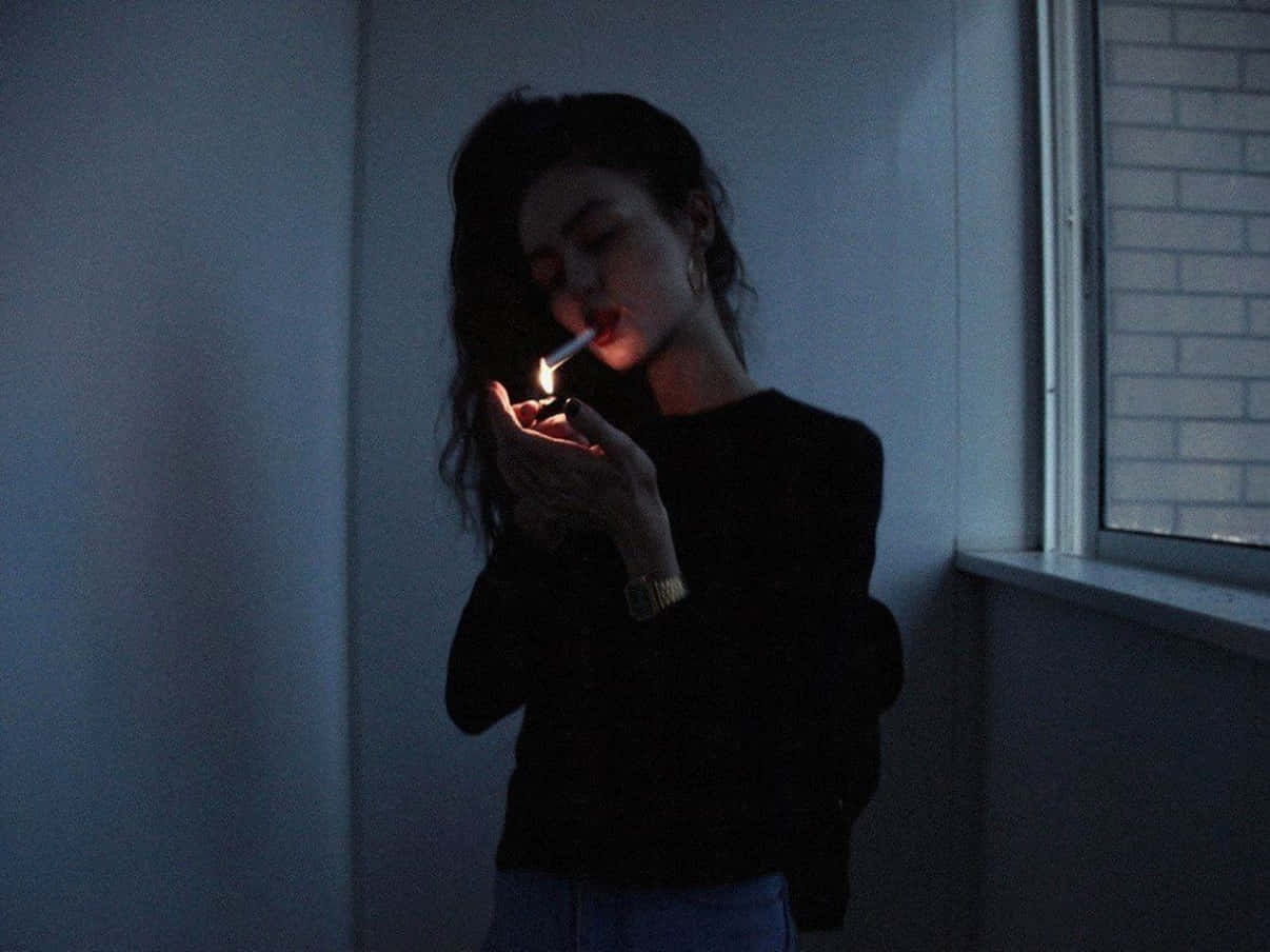 抽菸 桌布 壁紙 — 一個女人在昏暗的房間裡，在窗邊用打火機點燃一支香煙，溫暖的光芒映照在她的臉上。