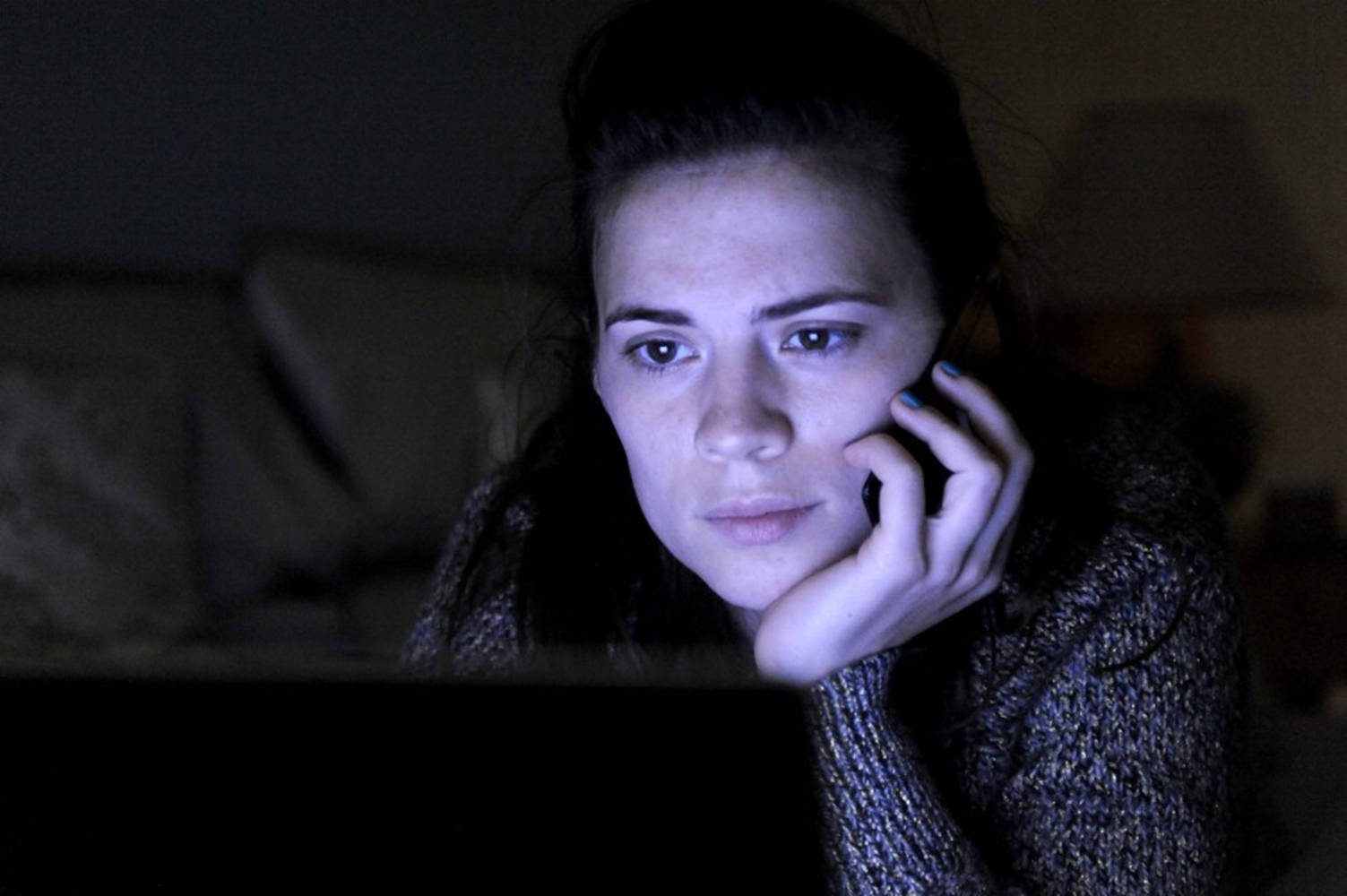 Netflix 桌布 壁紙 — 一個女人在昏暗的房間裡，帶著憂慮的表情專注地看著筆記型電腦螢幕，同時將電話貼在耳邊。