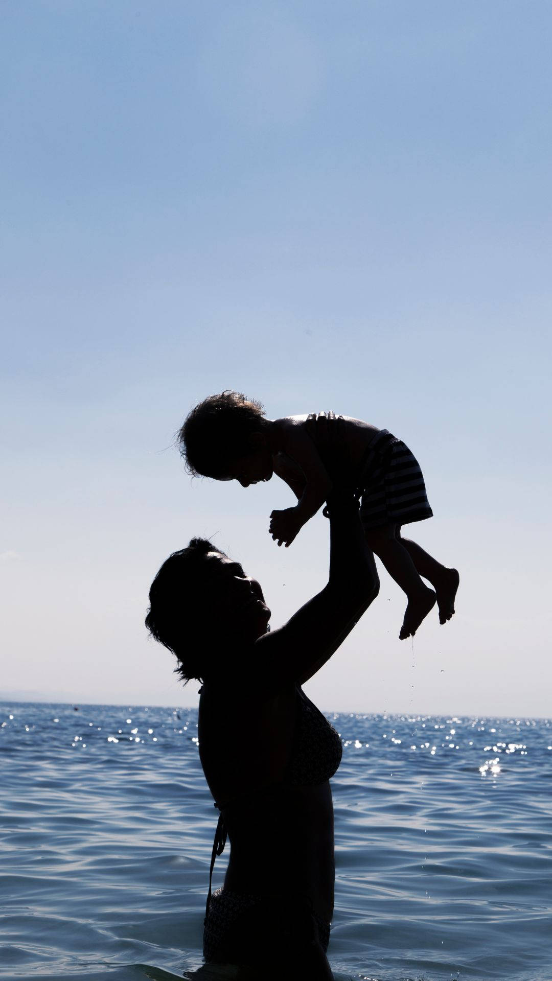 Mom 桌布 壁紙 — 一個女人在晴朗藍天下快樂地將孩子舉起在海浪之上的剪影。