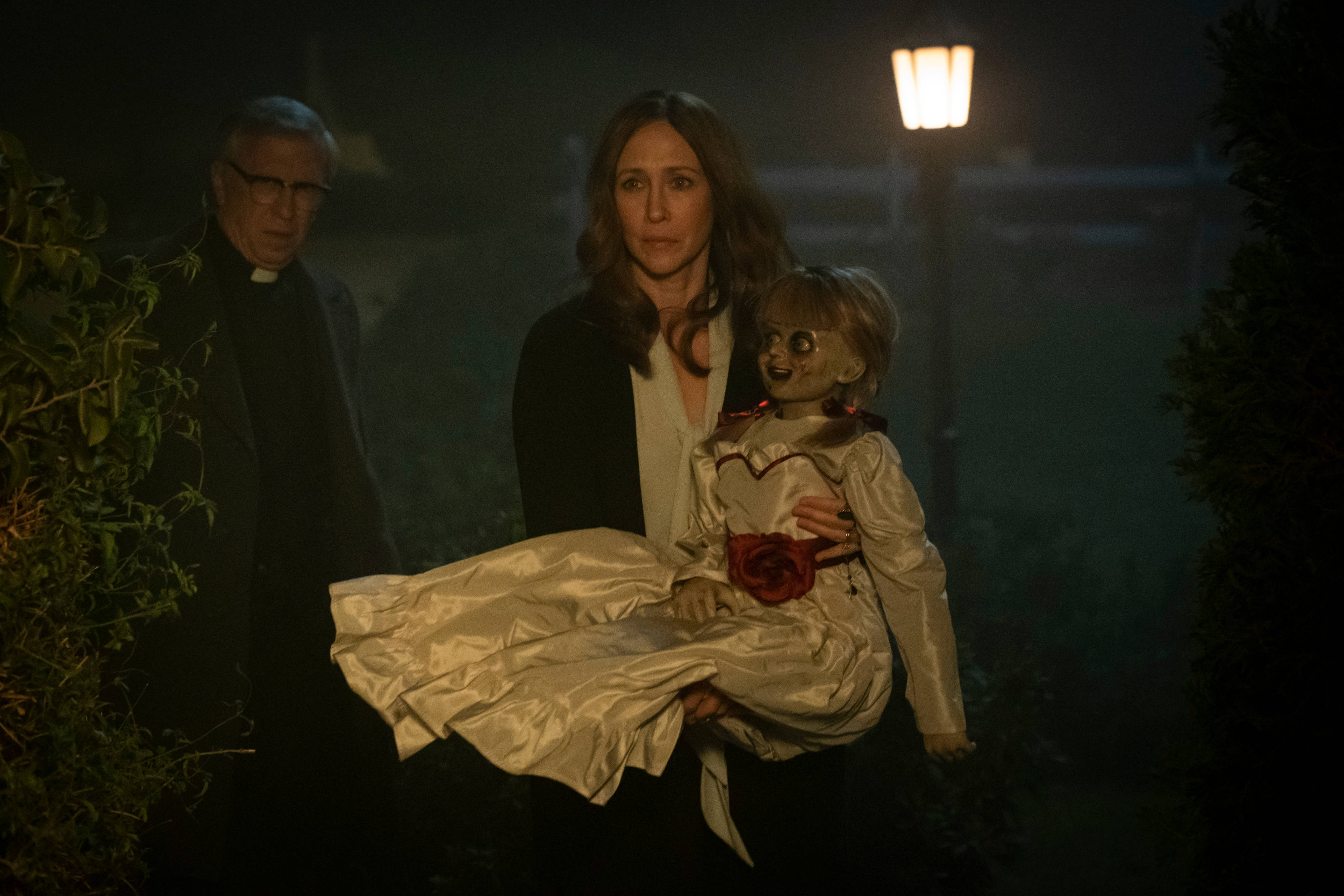 Annabelle 桌布 壁紙 — 一個女人在閃爍的路燈下拿著一個邪惡的娃娃，而一個牧師在黑暗的花園裡看著。