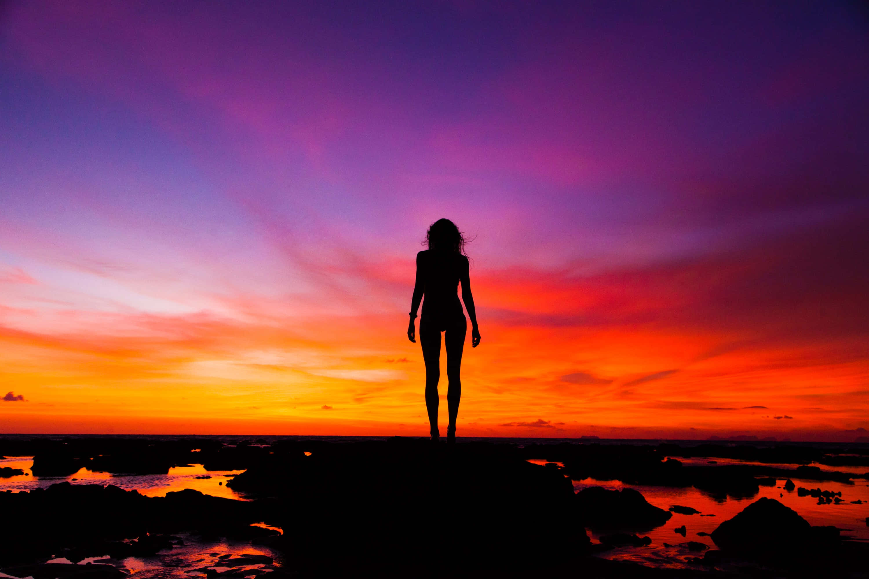 Silhouette 桌布 壁紙 — 一個女人獨自站在岩石海岸上的剪影，背景是紫色、橙色和紅色的火熱日落天空。