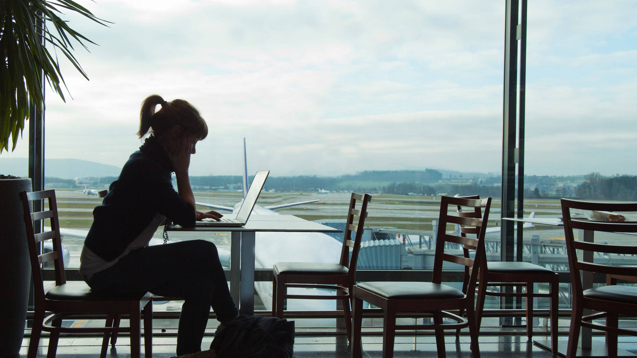 Airport 桌布 壁紙 — 一個女人的剪影，在窗邊用筆記型電腦工作，窗外是機場停機坪，有飛機和遠處的山丘，天空多雲。