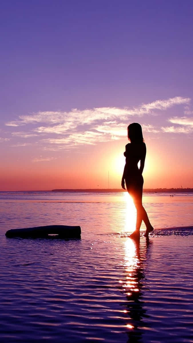 日落 桌布 壁紙 — 一個女人的剪影赤腳站在淺水中，夕陽在她身後落下，在漣漪的水面上投下金色的光芒。