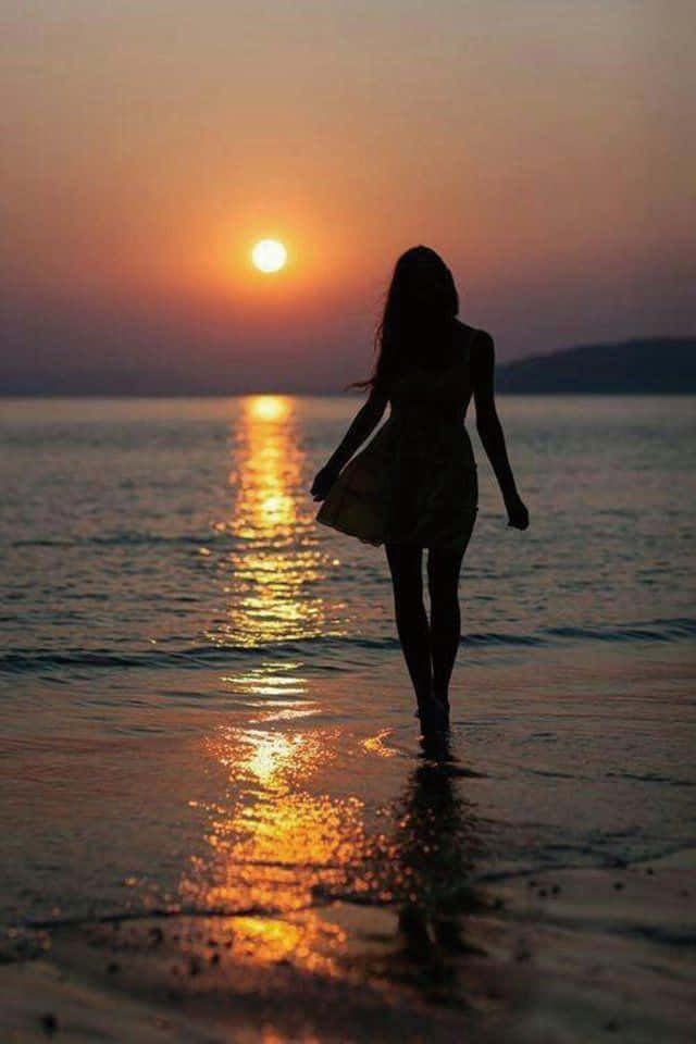 日落 桌布 壁紙 — 一個女人的剪影赤腳走在沙灘上，夕陽西下，在水面上投下金色的倒影。