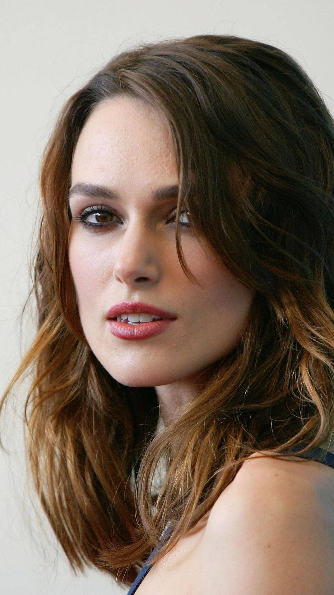 Keira Knightley 桌布 壁紙 — 一個女人的特寫肖像，她有波浪形棕色頭髮和柔和化妝，背景是純色。